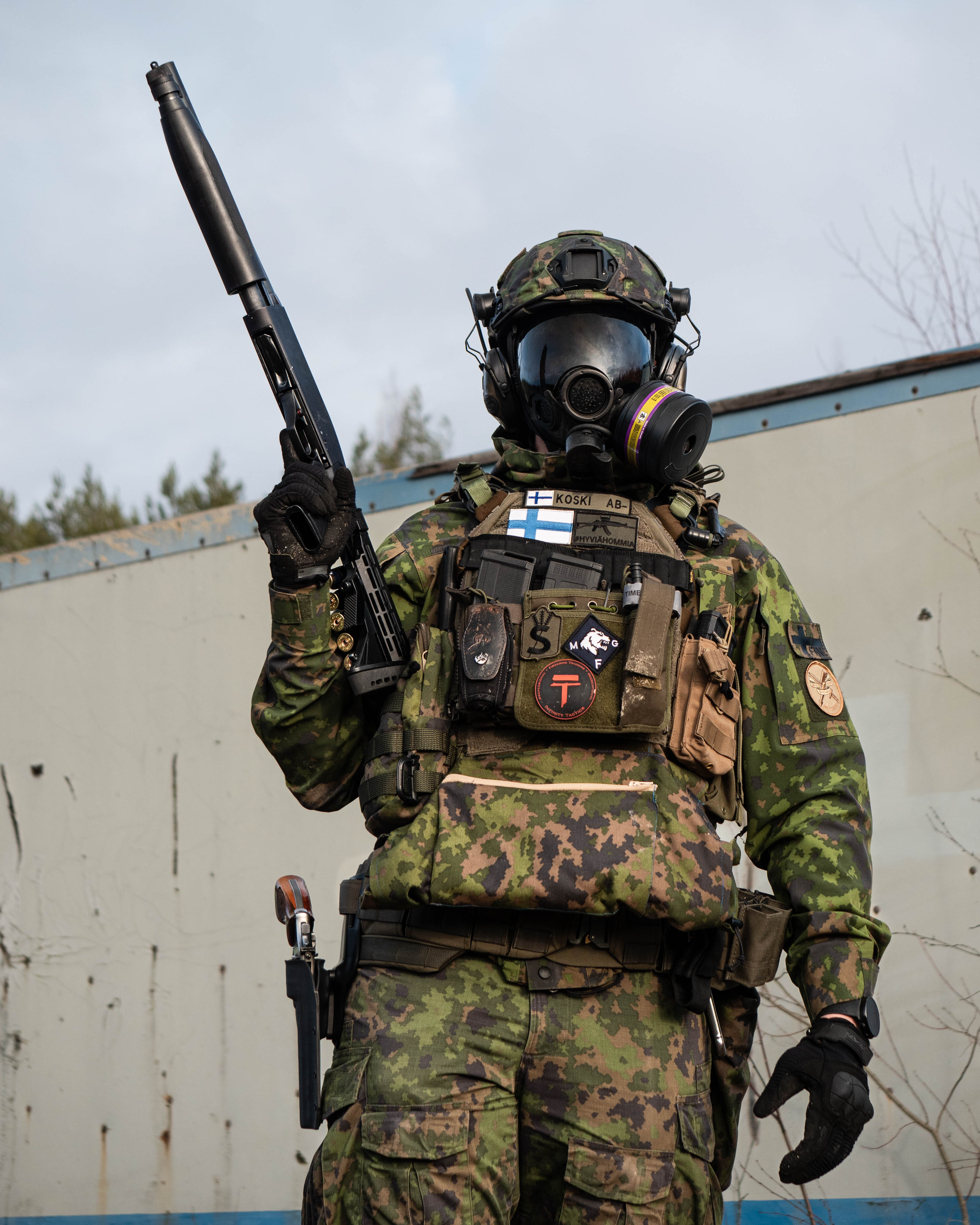 Man in army uniform and with a gas mask is standing infront of a container. He has a shotgun.