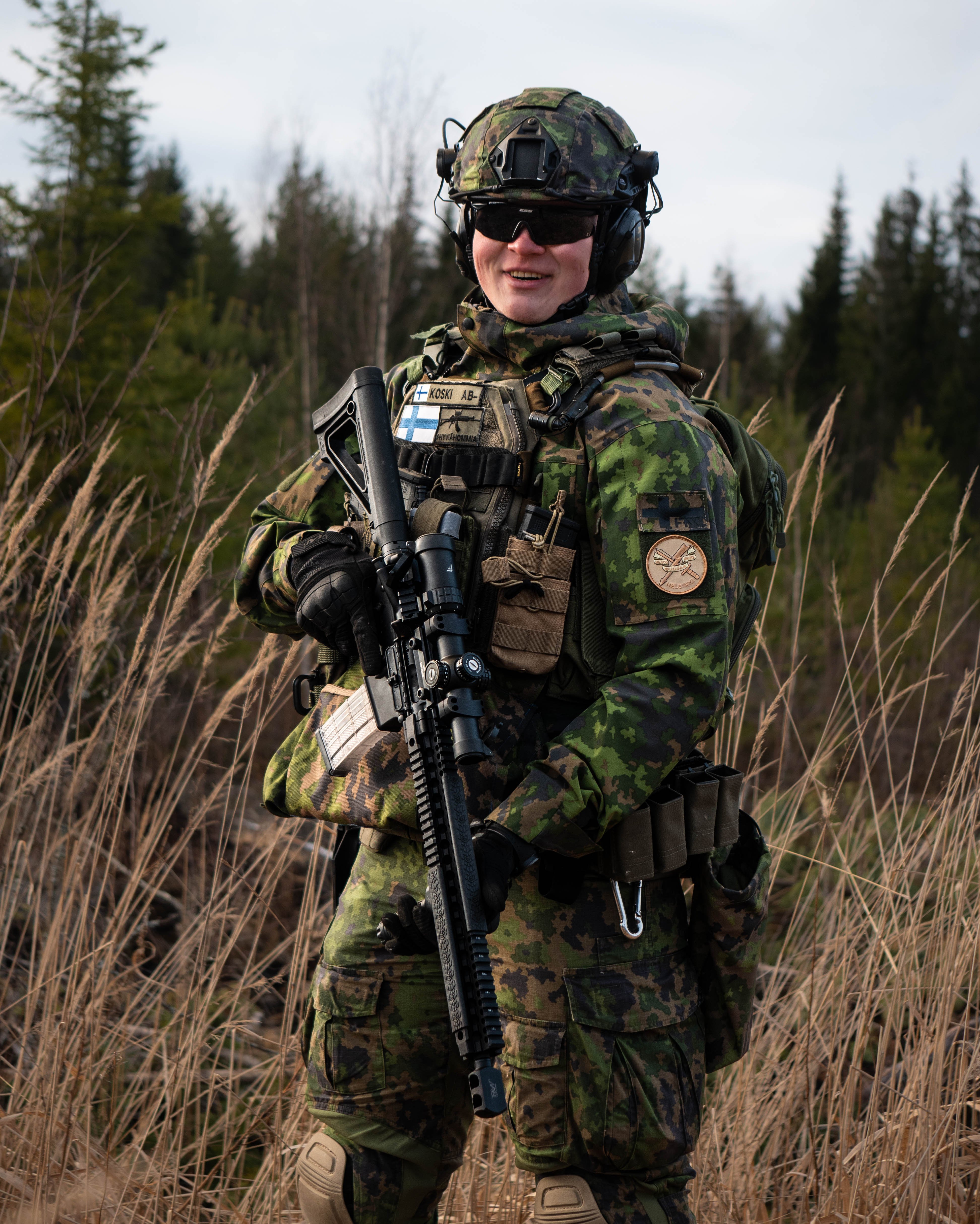 Man in army uniform is standing in the middle of a field. He has a rifle.