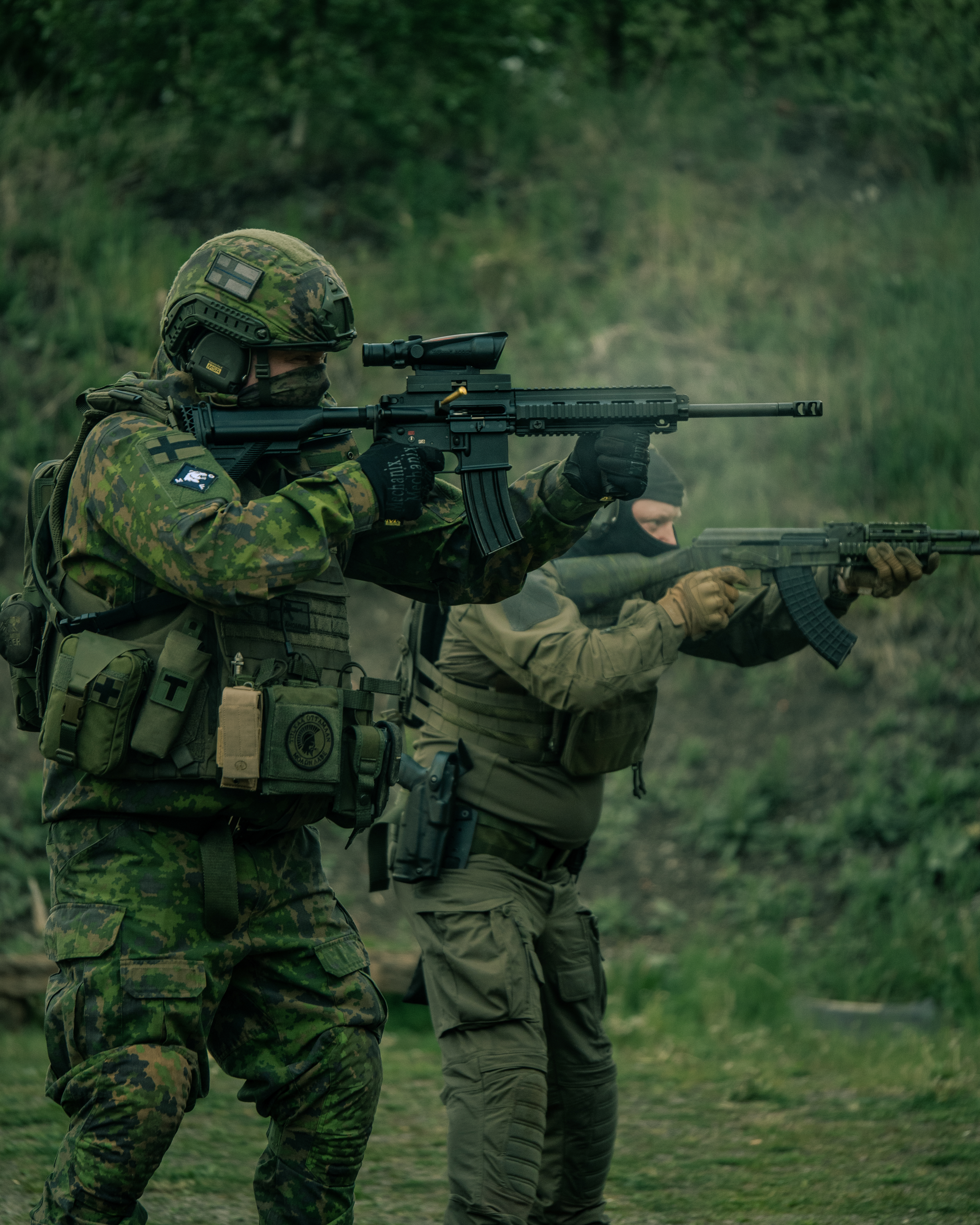 Two men with rifles in a high stance are shooting on a shooting range. One of men has a ak47 rifle, he is wearing a M05 camo jacket, M05 camo pants, a plate carrier, boots and a protection group denmark pgd ballistic helmet. The other man is wearing a balaclava, earpro, a uf pro combat shirt, uf pro tactical pants, boots and a gun belt. He has a M4 AR15 Heckler & Koch 416 with a ACOG optic. Shell casings are flying through the air.  Finnish reservists