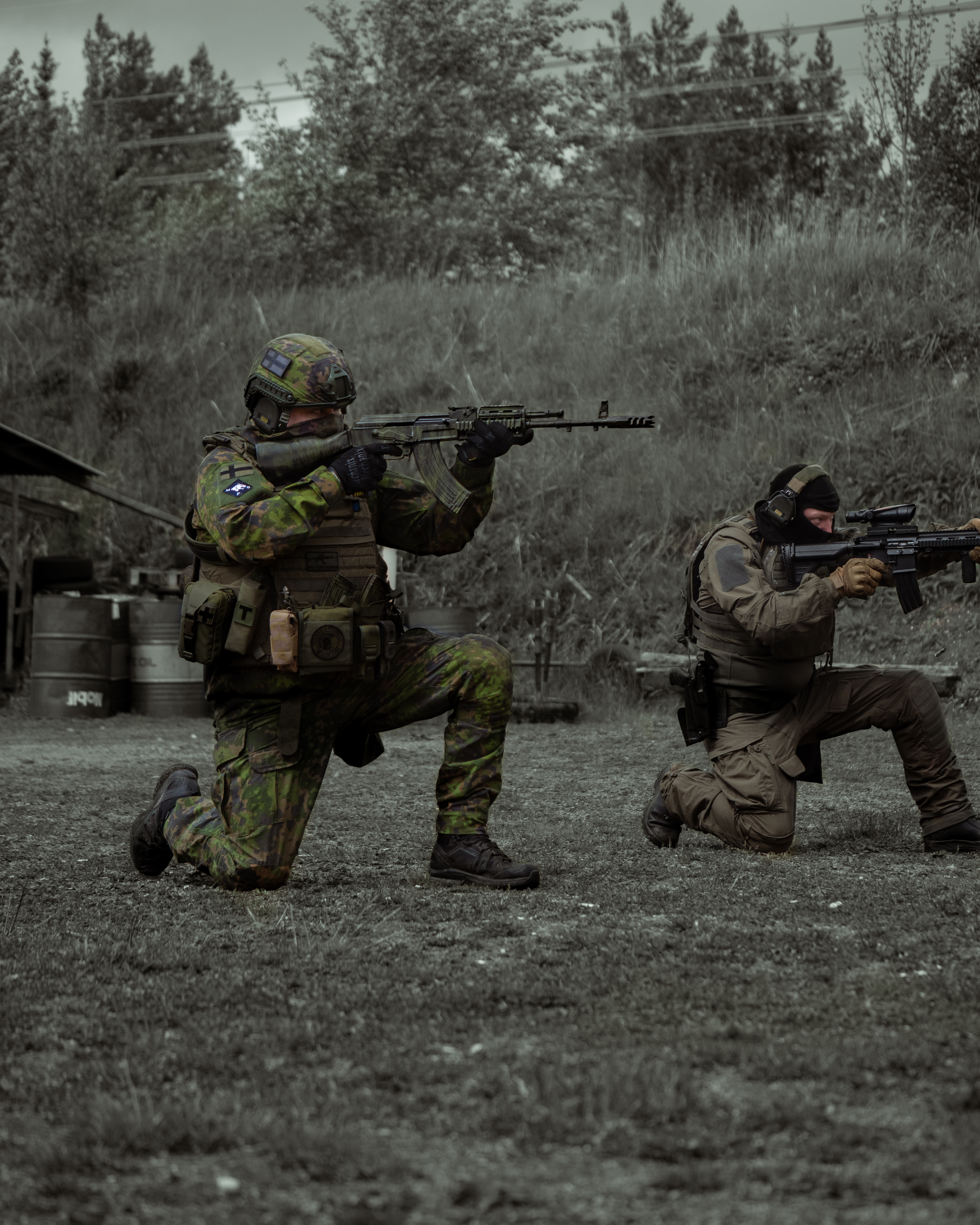 Two men with rifles in a high knee stance are shooting on a shooting range. One of men has a ak47 rifle, he is wearing a M05 camo jacket, M05 camo pants, a plate carrier, boots and a protection group denmark pgd ballistic helmet. The other man is wearing a balaclava, earpro, a uf pro combat shirt, uf pro tactical pants, boots and a gun belt. He has a M4 AR15 Heckler & Koch 416 with a ACOG optic.  Finnish reservists
