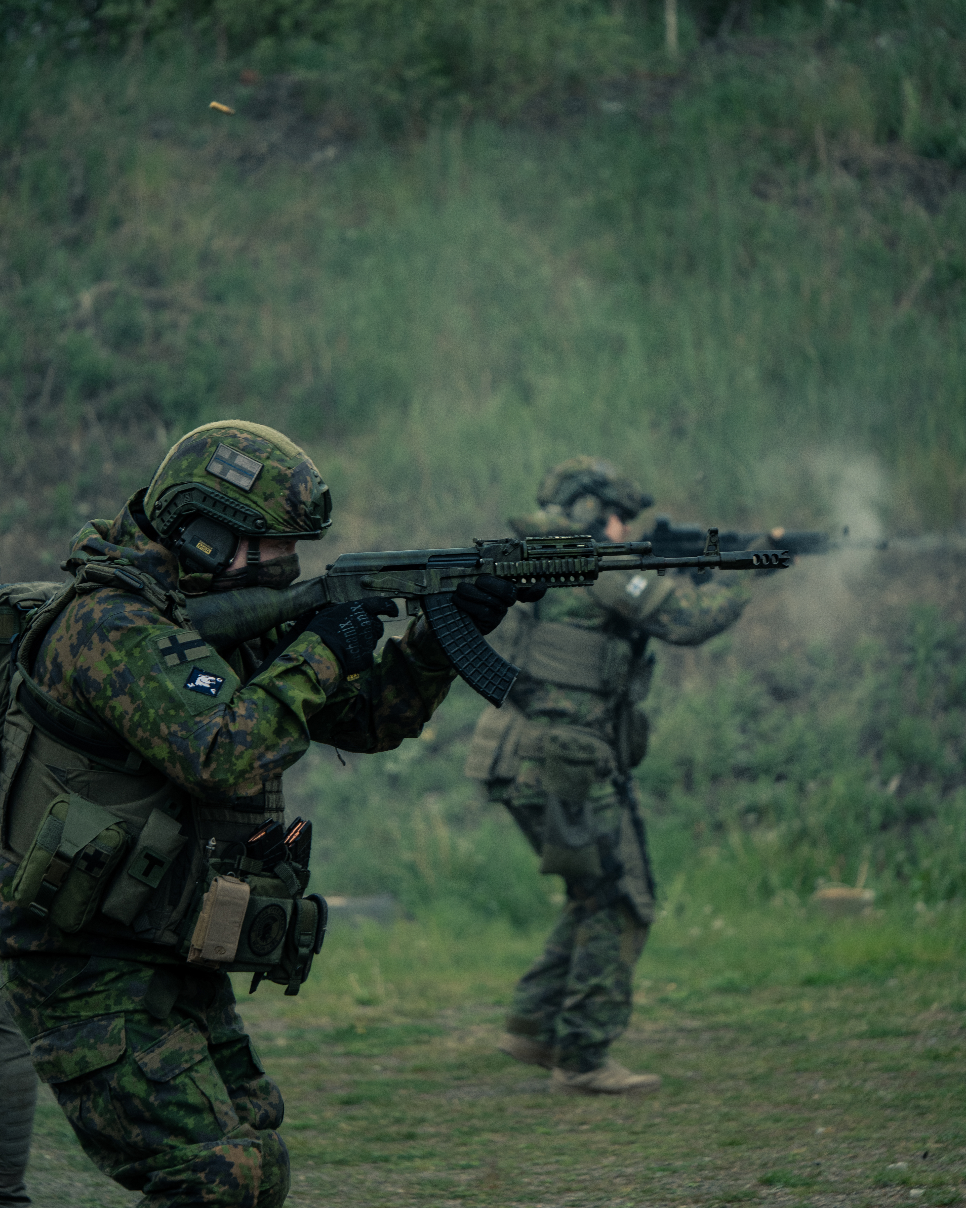 Two men with rifles in a high stance are shooting on a shooting range. One of men has a ak47 rifle, he is wearing a M05 camo jacket, M05 camo pants, a plate carrier, boots and a protection group denmark pgd ballistic helmet. The other man is wearing a M05 clothing, a plate carrier, a ballistic helmet and a  battle belt. He has a ak-47 rifle with a eotech holographic sight. Shell casings are flying through the air.  Finnish reservists