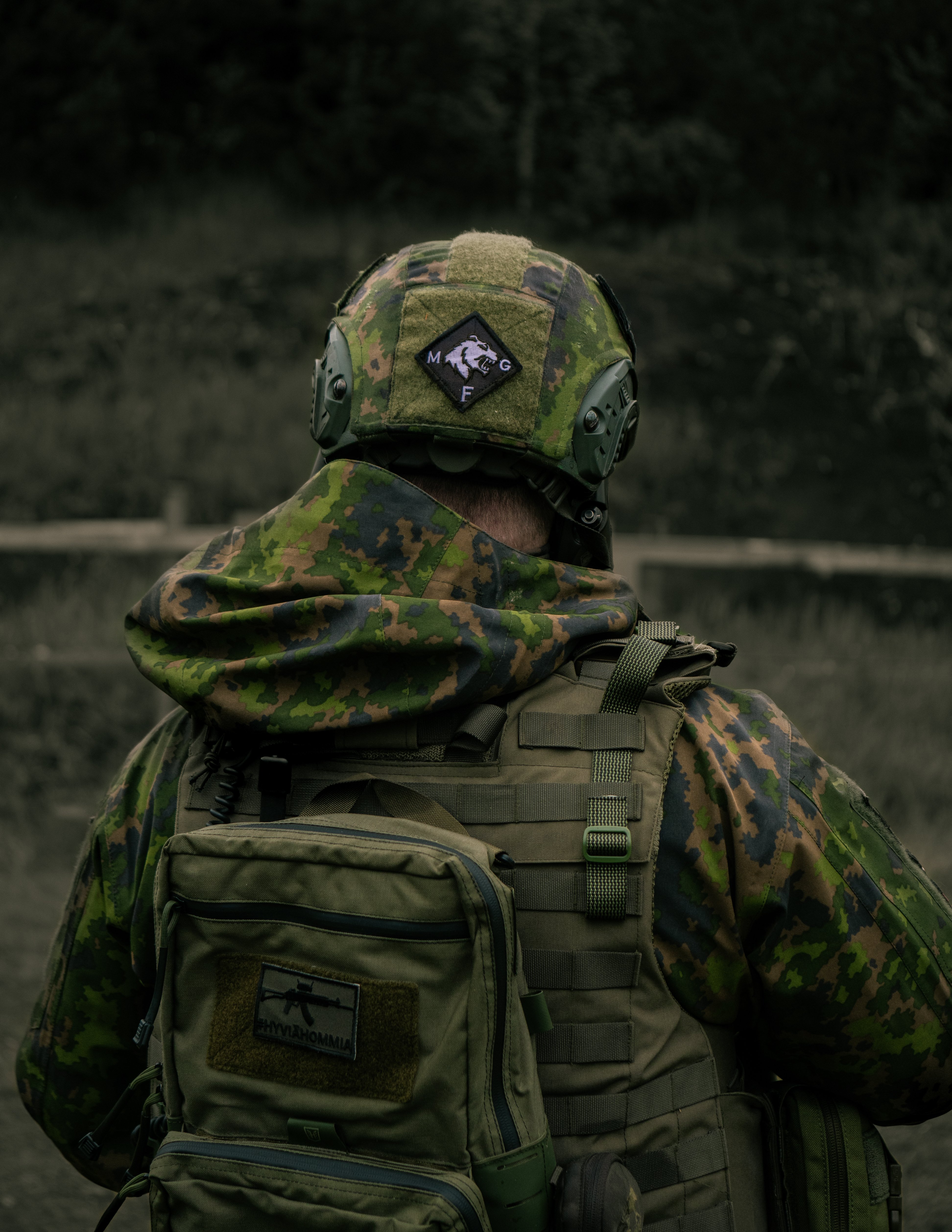 A man with his back turned to the camera is standing on a gun range. He is wearing a balistic helmet with a M05 camo cover and a MGF military group finland patch. He is also wearing a a M05 field jacket with a hoodie and a plate carrier and a #hyviähommia patch. Finnish reservist