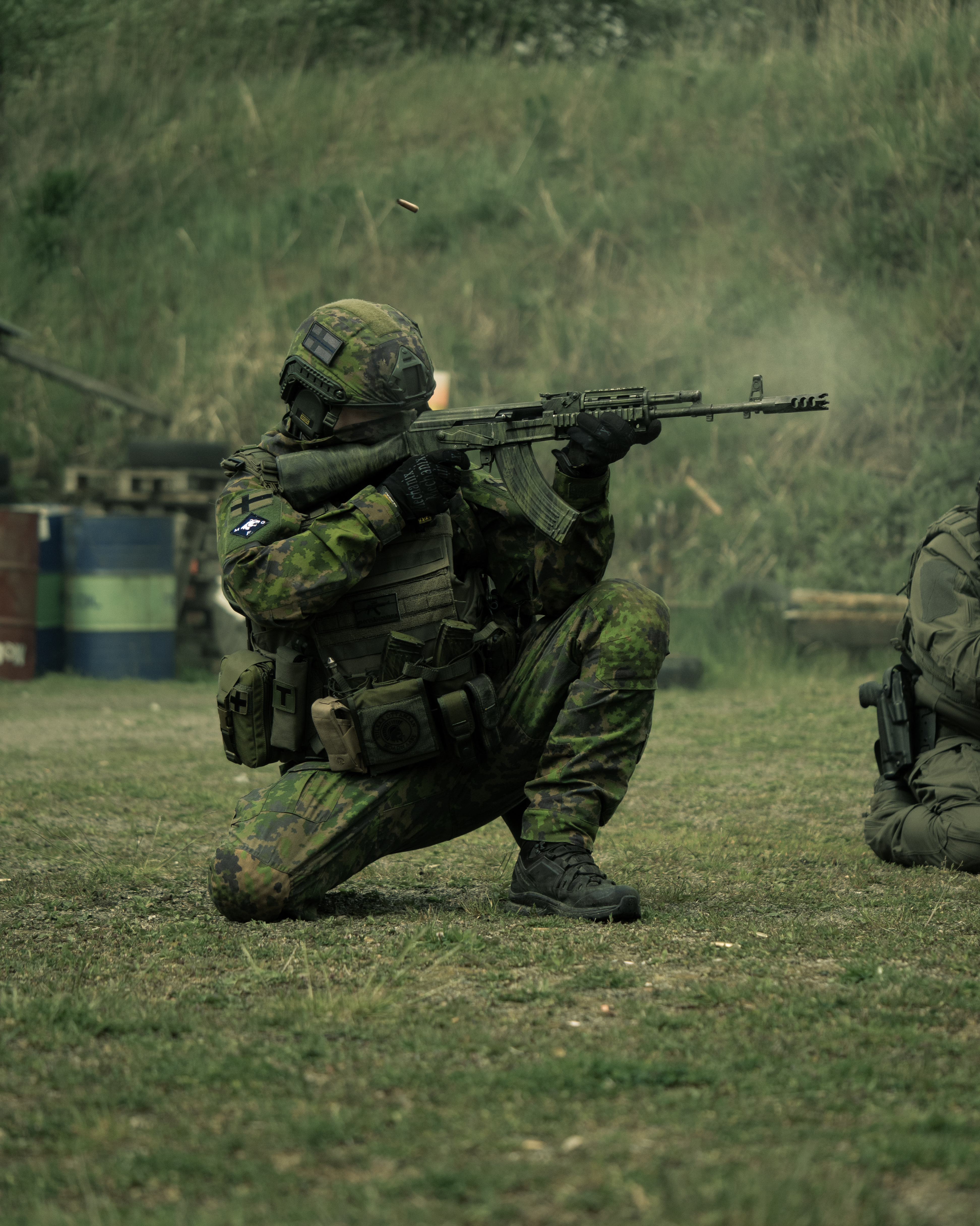 Man is kneeling with rifle on a gun range. Finnish Reservist. He is wearing a M05 camo jacket and M05 combat pants. He has a ballistic helmet. He has a ak-47 rifle. He has a Finnish flag patch and other patches on his arm.