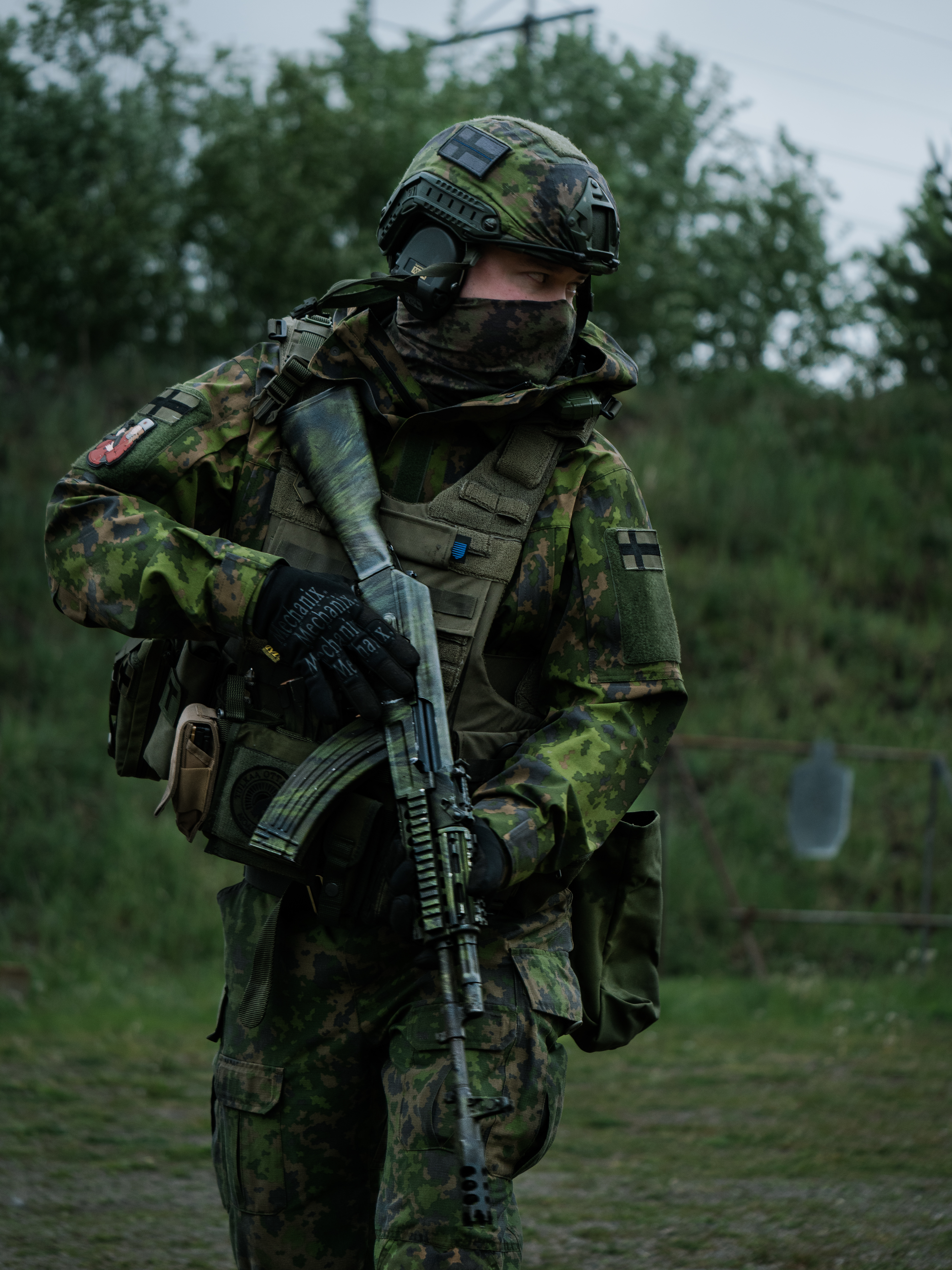 Man is turning around with rifle. Reservist. He is wearing a M05 camo jacket and M05 combat pants. He has a ballistic helmet. He has a ak-47 rifle. He is on a shooting range. He has a Finnish flag and other patches on his arm.