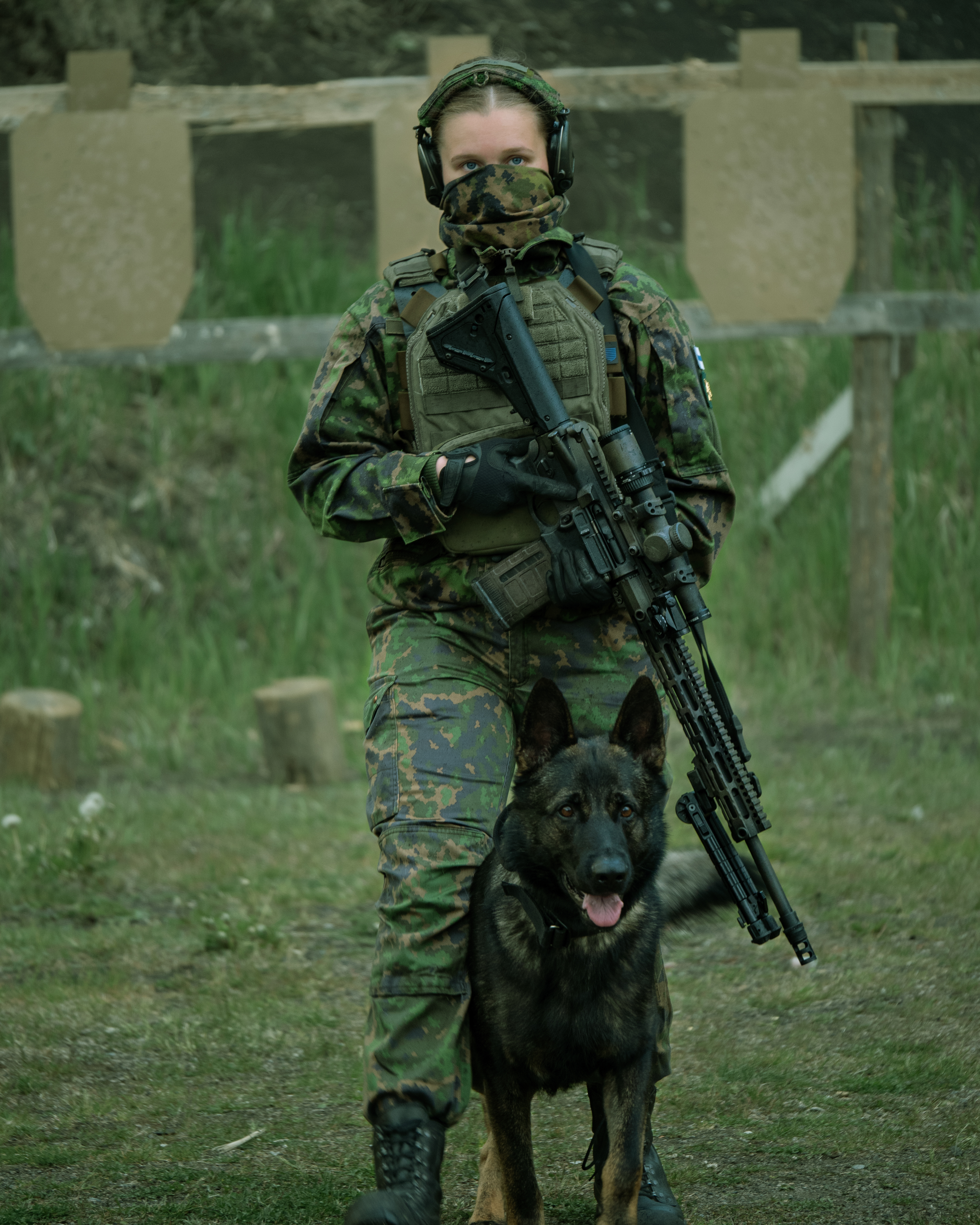  A woman in army cloathing is standing on a sshooting range with a dog K9. She is holding a 308. AR10 rifle with a optic. She is wearing a facemask, earphones, a  M05 camo jacket, M05 camo pants and combat boots.  Female finnish reservist.