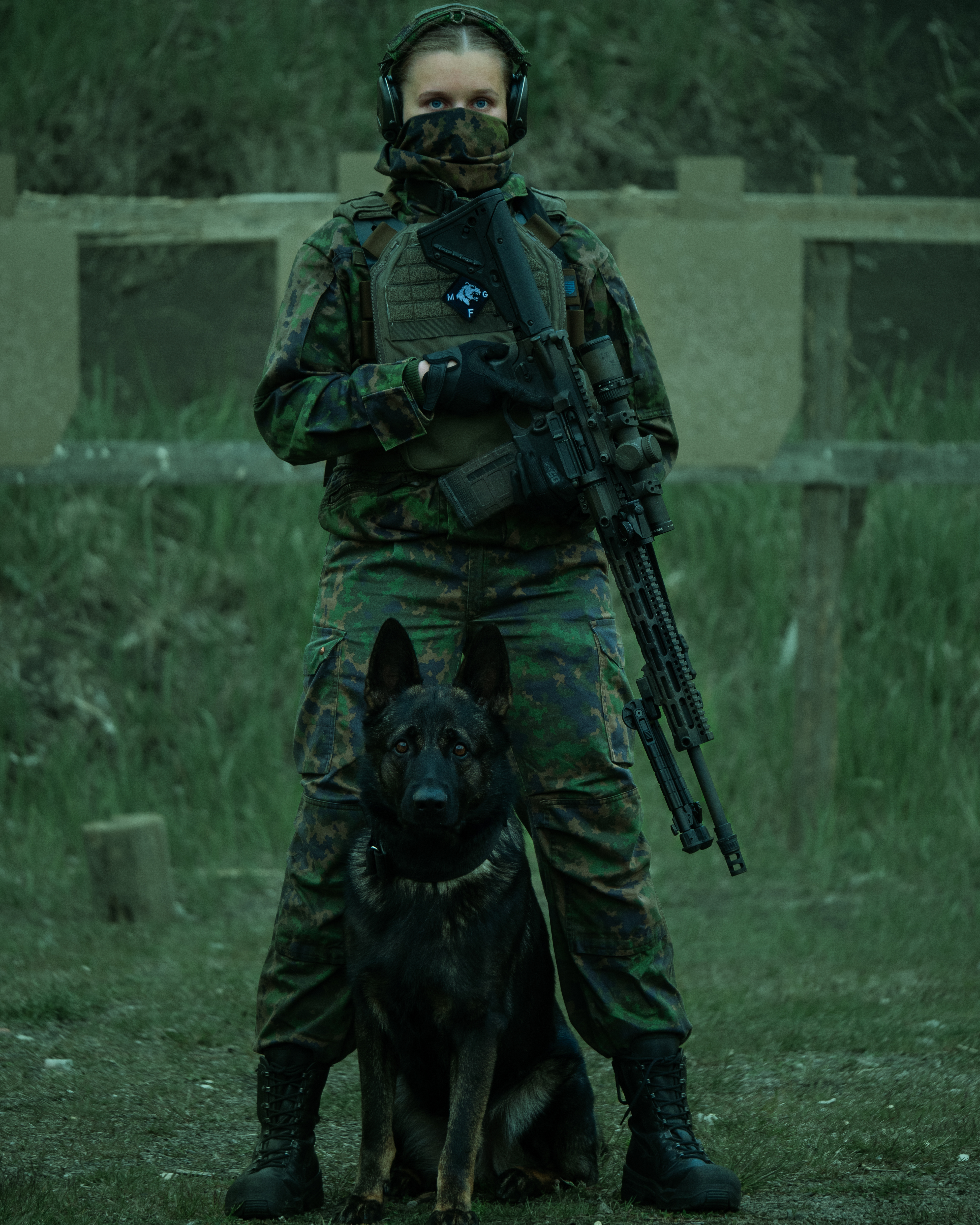 A woman in army cloathing is standing on a sshooting range with a dog K9. She is holding a 308. AR10 rifle with a optic. She is wearing a facemask, earphones, a  M05 camo jacket, M05 camo pants and combat boots.  Female finnish reservist.