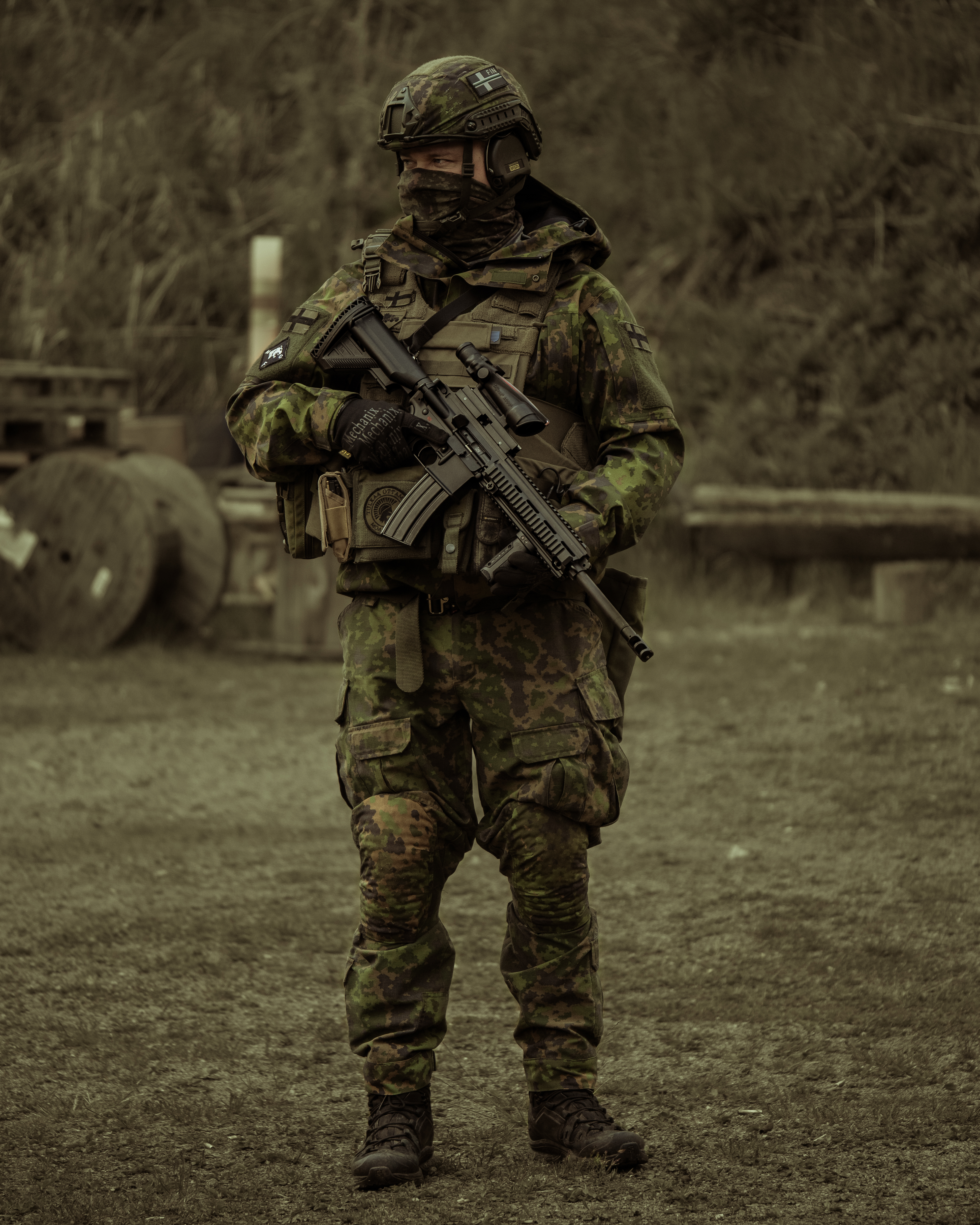 A man is standing around on a gun range. He has a M4 AR15 Heckler & Koch 416 with a ACOG optic, he is wearing a M05 camo jacket, M05 camo pants, a plate carrier, boots and a protection group denmark pgd ballistic helmet. Finnish reservists