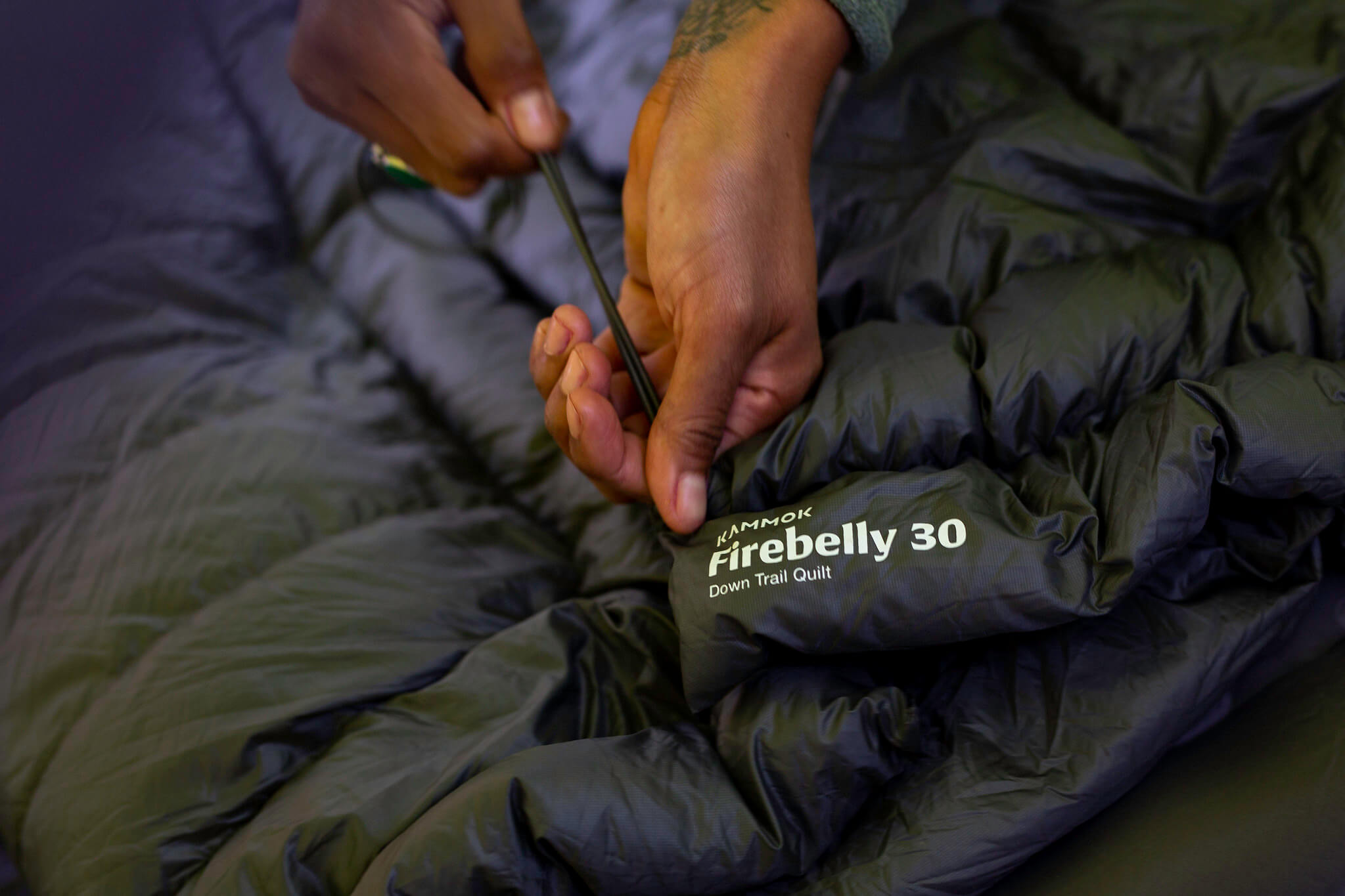 Hands cinching the adjustable shock cords on corners of a Kammok Firebelly 30 trail quilt in Granite Gray.