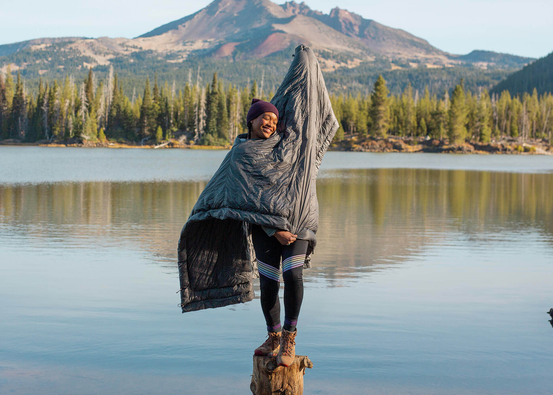 Kammok Firebelly 30 poncho quilt in Granite Gray, worn in hands-free poncho mode by girl standing on log in shallow water.