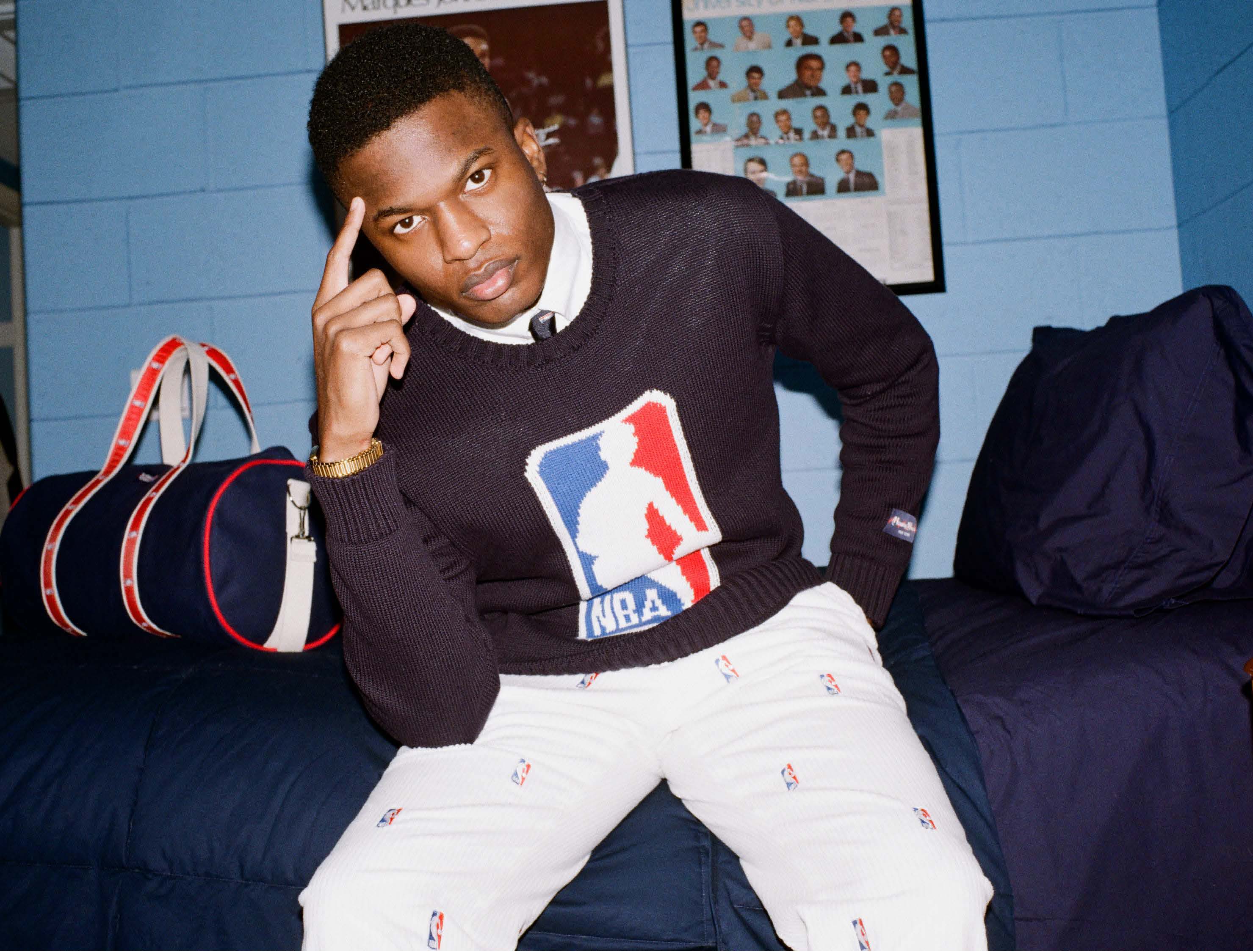 Model wearing the NBA Logo Sweater in Navy on a bed next to the NBA Logo Banker Bag.