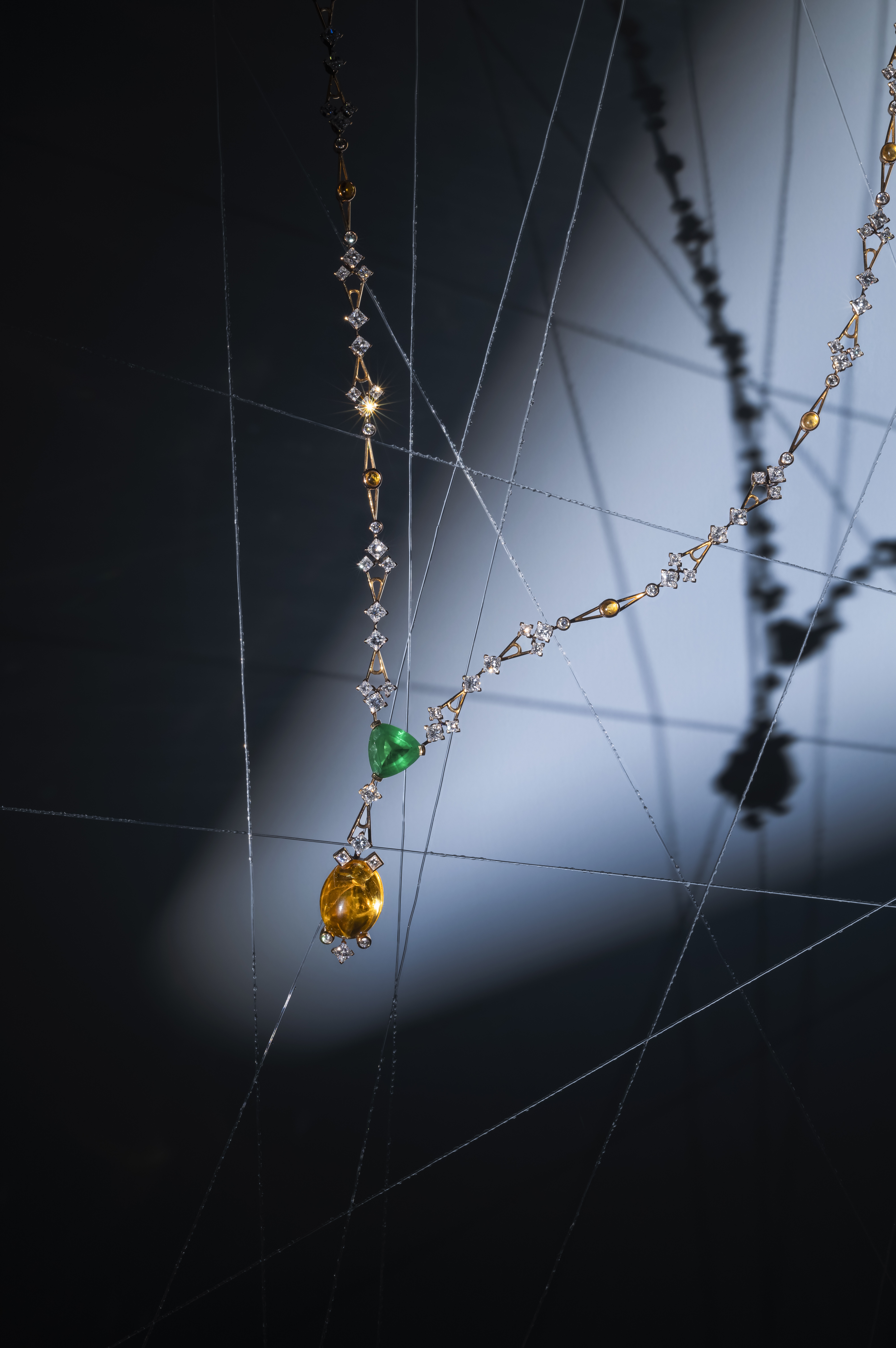 A delicate necklace with a silver chain, featuring sparkling diamonds, a yellow gemstone pendant, and an emerald gemstone. The necklace is artistically displayed against a dark, blurred background with intersecting thin lines, resembling a spiders web.
