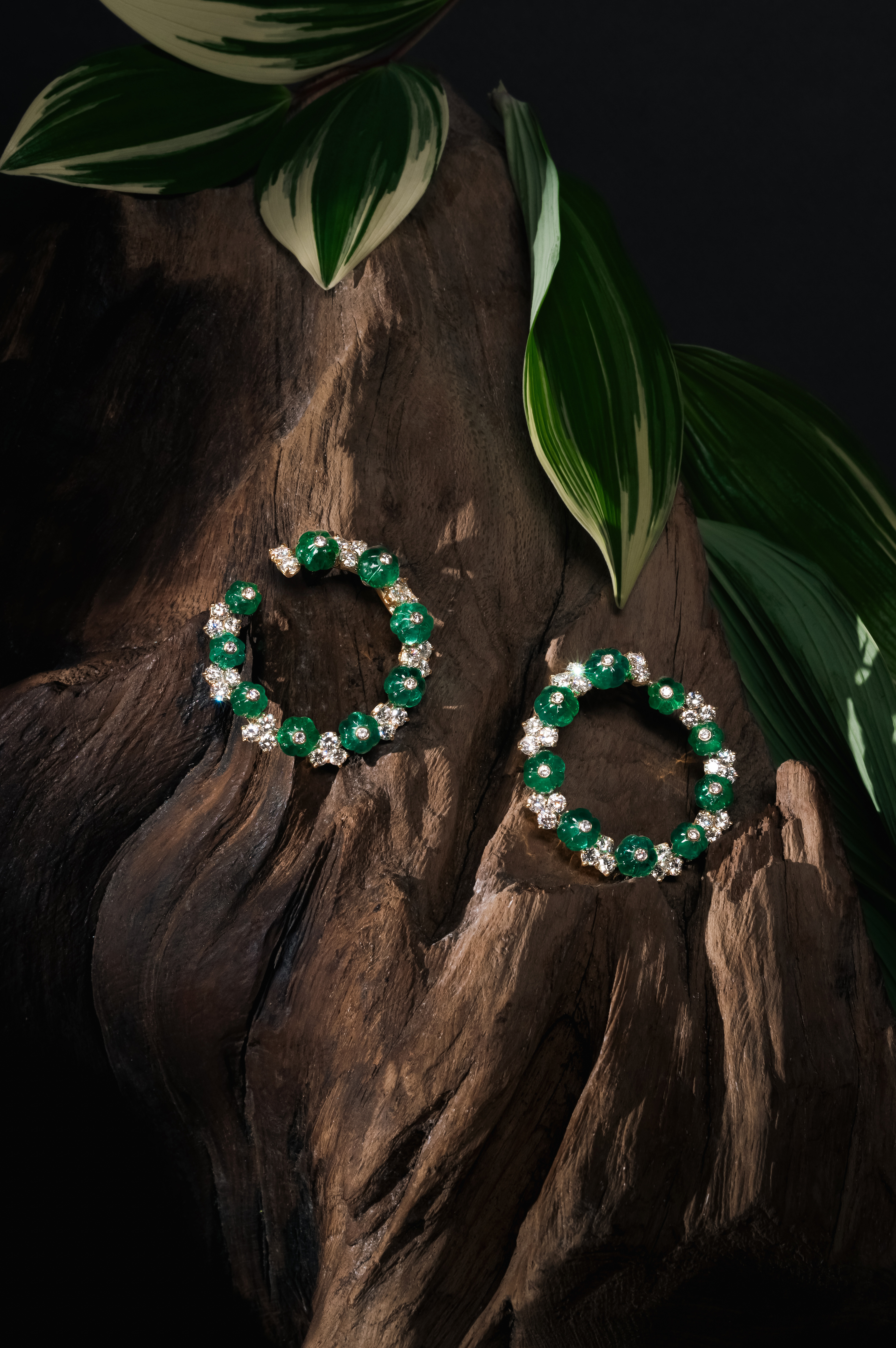 A pair of circular emerald and diamond earrings are elegantly displayed on a piece of dark, textured wood, adorned with green foliage. The background is black, highlighting the rich colors of the gemstones and the natural beauty of the display elements.
