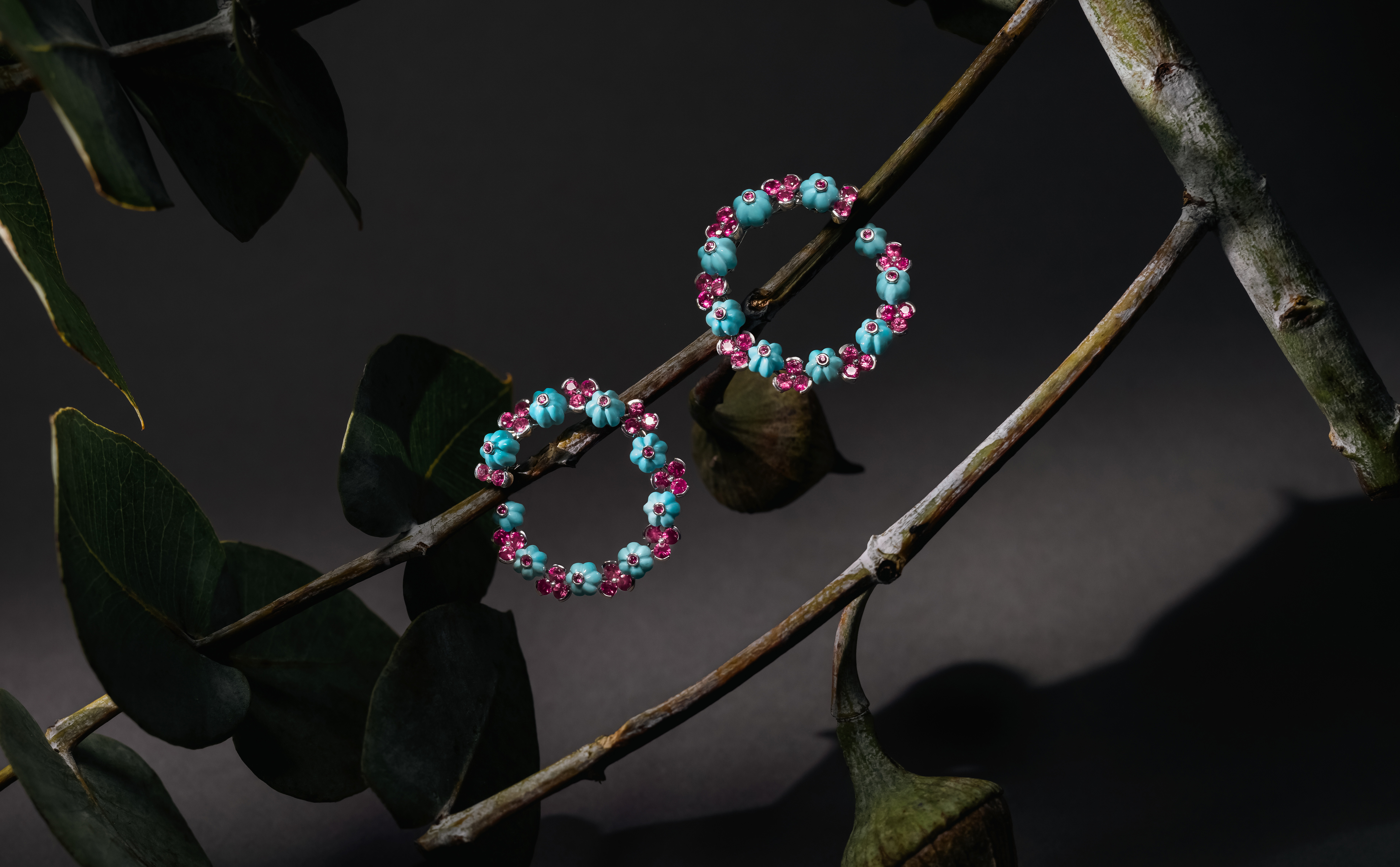 A pair of colorful beaded hoop earrings is displayed on a branch with dark green leaves. The earrings feature small blue and pink beads arranged in a circular pattern, set against a dark black background.