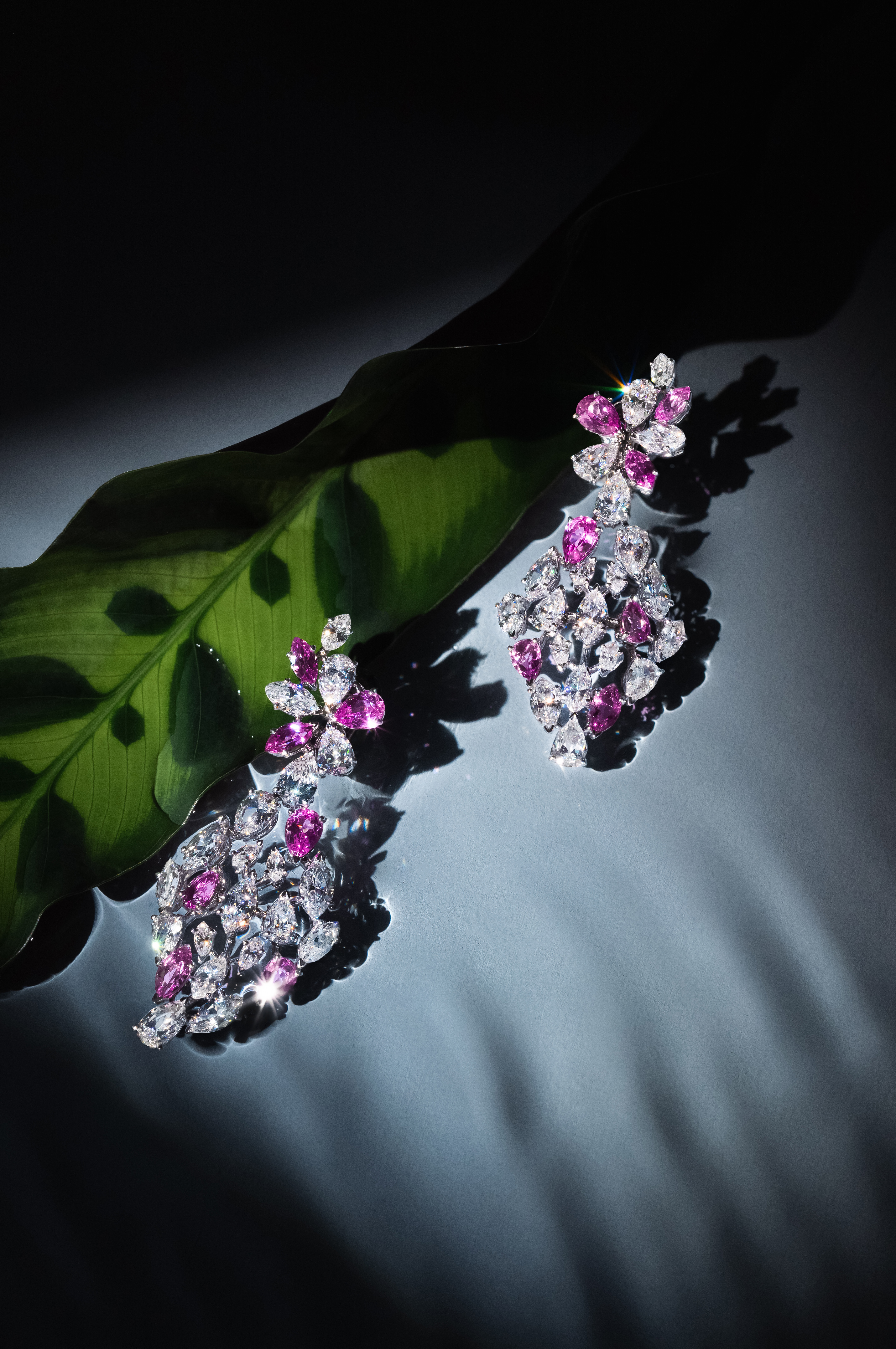 A pair of elegant floral-shaped earrings adorned with pink and white gemstones displayed against a dark background with a green leaf highlighting the intricate details. The lighting focuses on the jewelry, creating a dramatic and luxurious effect.
