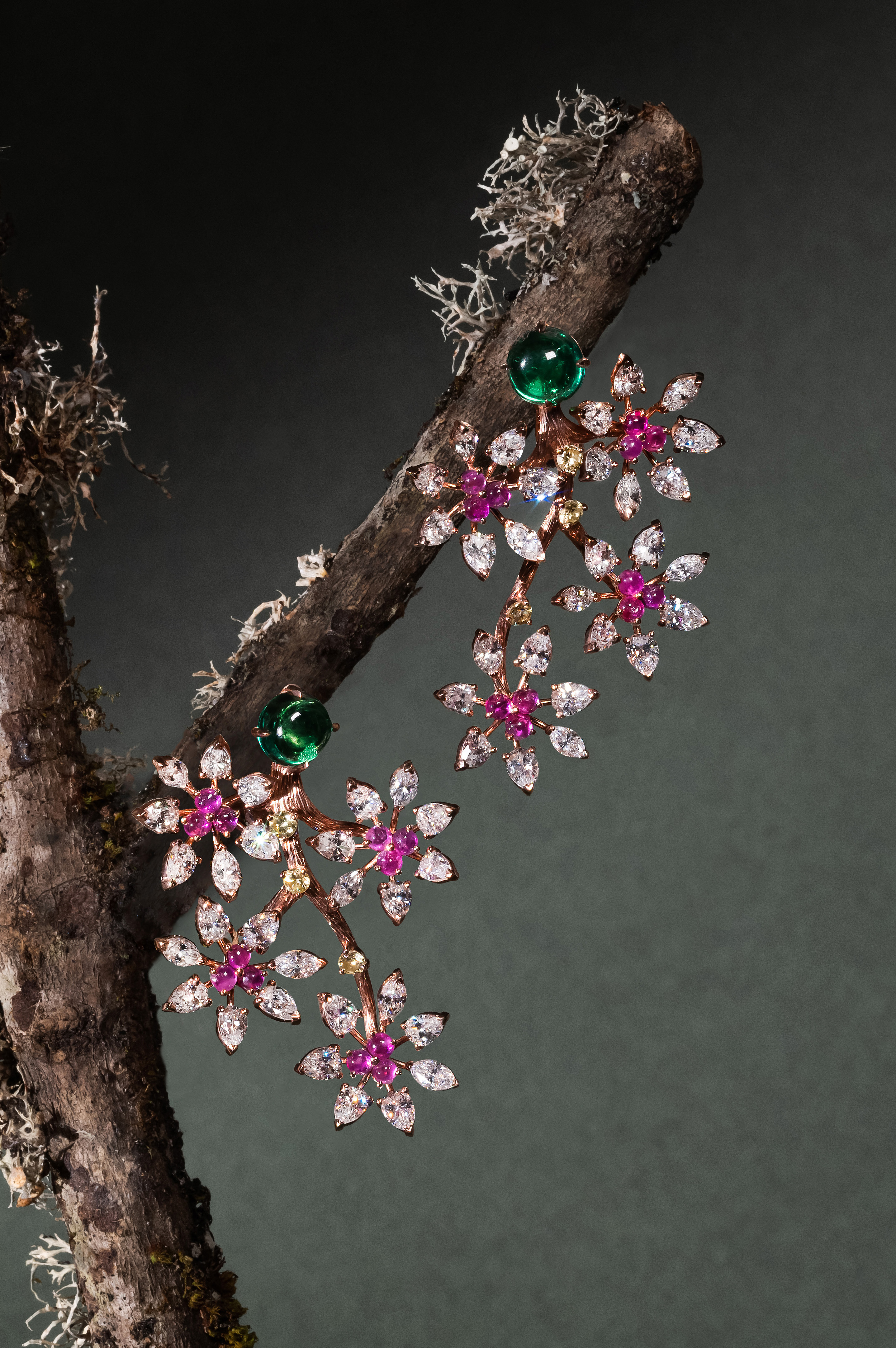 A pair of intricate, colorful earrings is draped over a rough, bare tree branch. The earrings feature a cluster of small, sparkling gemstones, including pink and green stones, arranged in a floral pattern. The background is dark, adding contrast to the bright jewelry.
