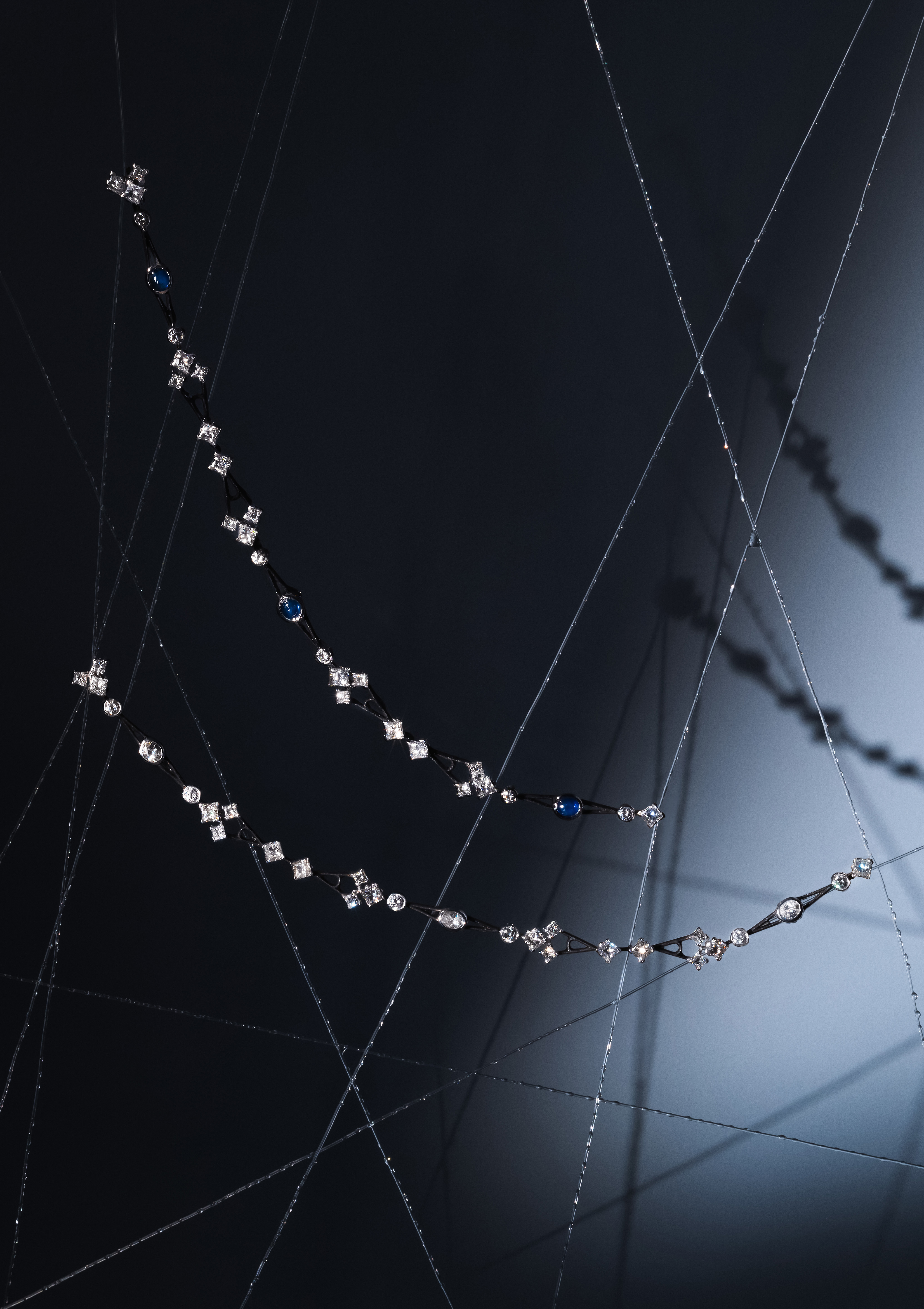Delicate, white gold bracelets with variously shaped diamonds and dark gemstones are artistically displayed against a black and white gradient background with reflective elements. Thin, intersecting wires add an abstract and sophisticated touch to the composition, resembling a spiders web.