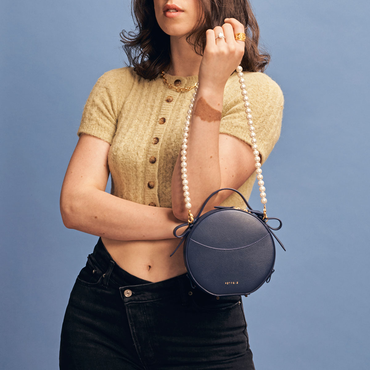 Circa Bag Pebbled Marine with Gold Hardware Pearl Shoulder Chain Attached Woman with Fair Skin Holding the Bag in Front of Her Wearing a Yellow Collared Shirt and Black Pants