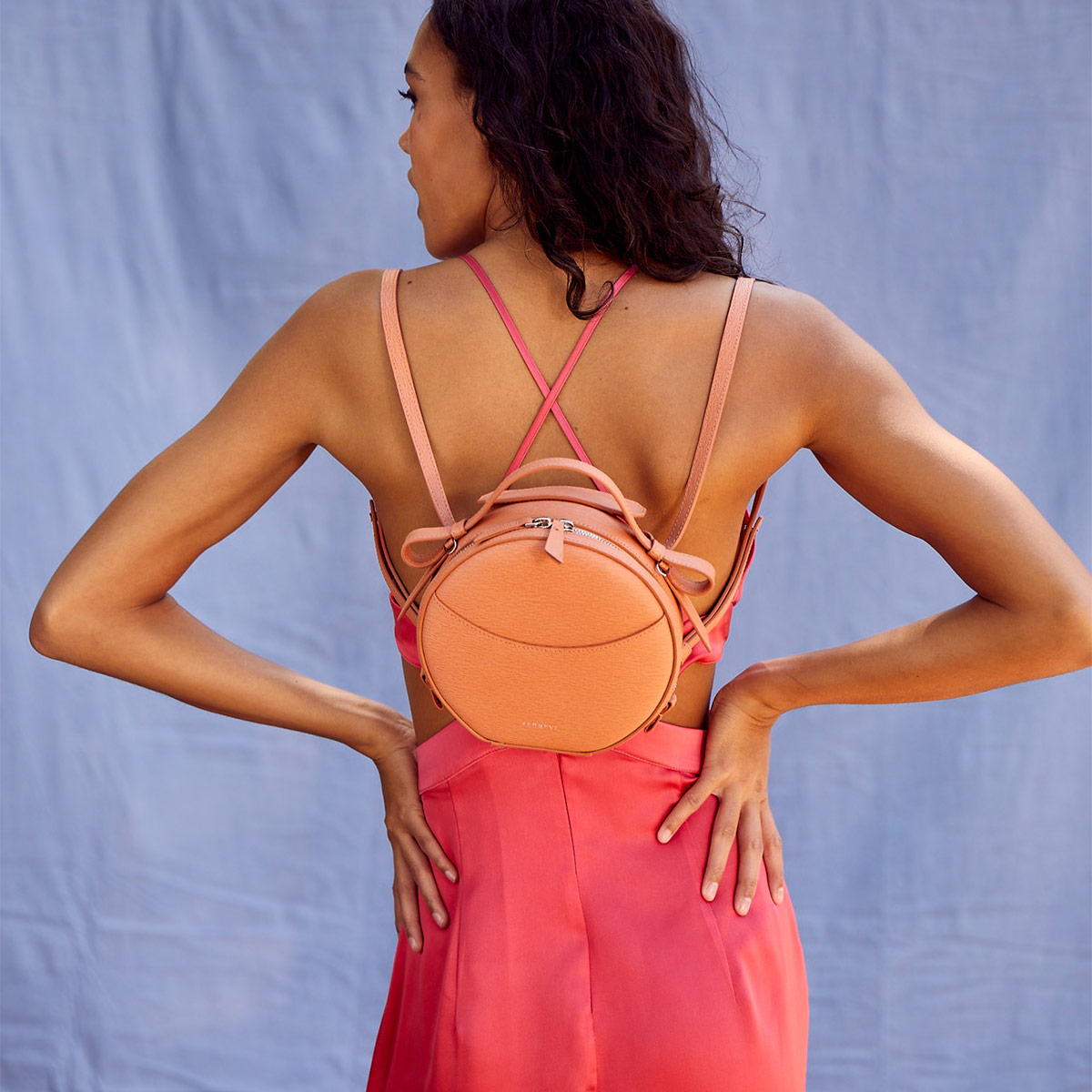 model against a blue background wearing a pink dress and wearing a pink circle bag as a backpack