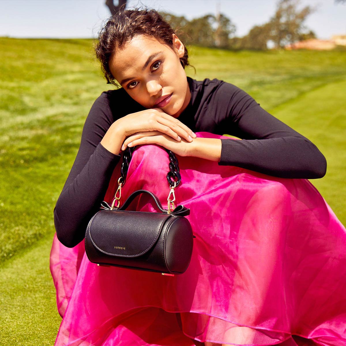 Model with Light Brown Skin and Hair Pulled Into a Low Bun. The model is wearing a black turtleneck and a bright pink flowy and silky skirt. She has a Mini Barrel Bag Pebbled Noir, Flat Acetate Chain in Noir Attached propped upon her knee.