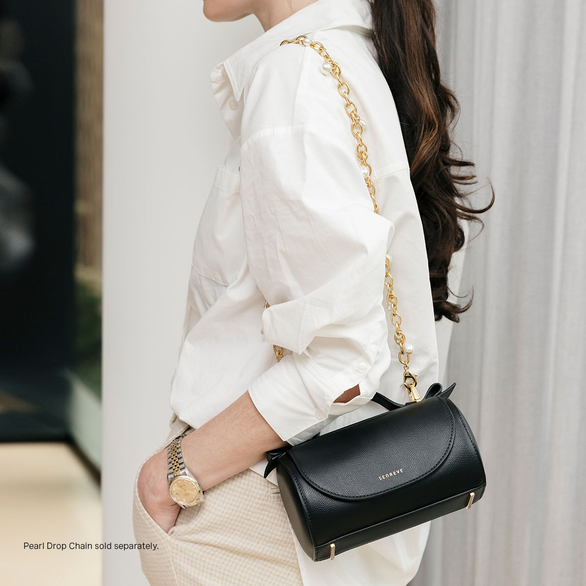 Side View of Mini Barrel Bag Pebbled Noir with Gold Beveled Shoulder Chain Strap Attached. Bag is on the shoulder of a white woman wearing a gold watch, white business shirt, and tan khakis. 