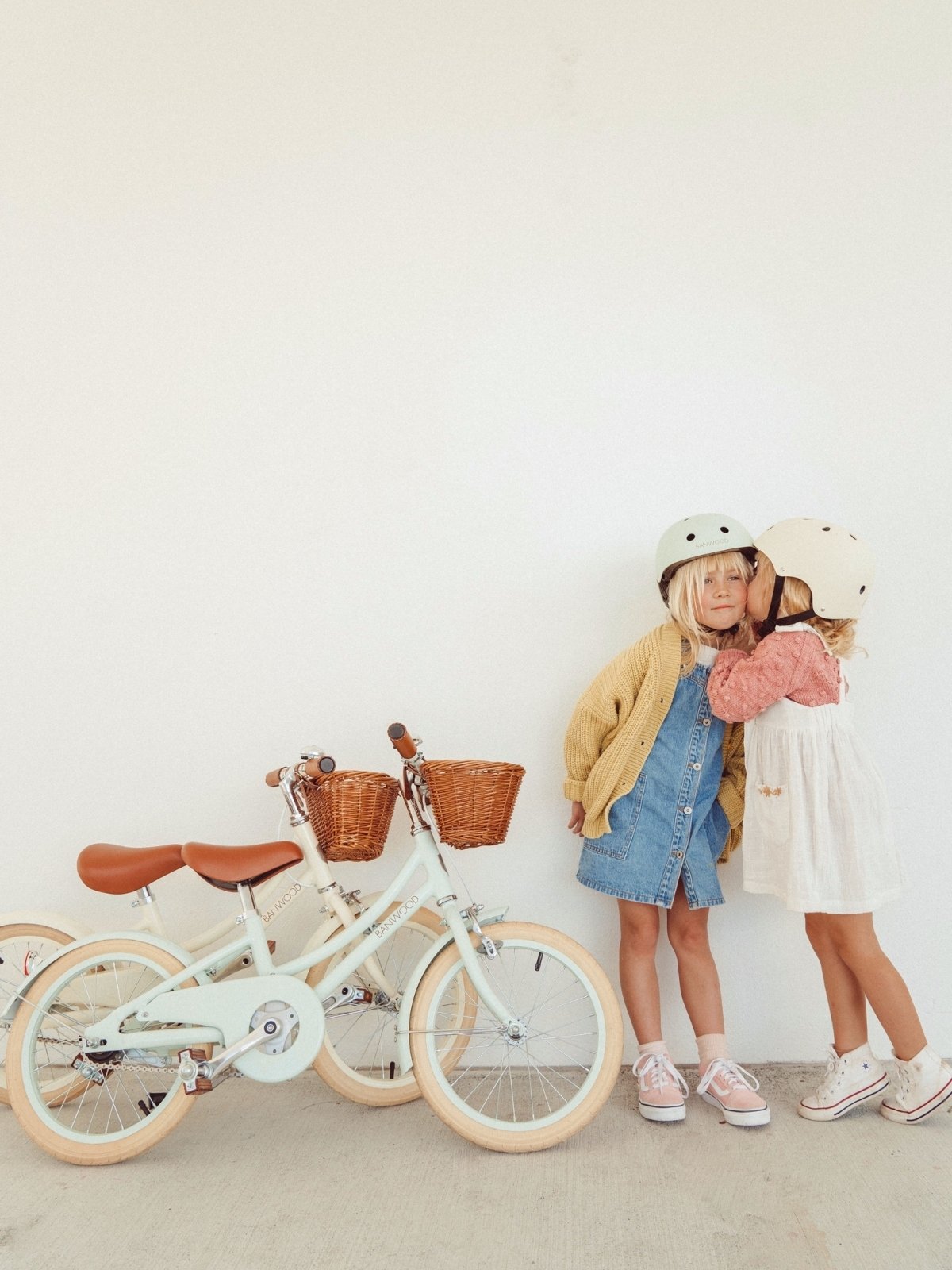 girl whispering into another girls ear, standing next to two banwood bikes, one is blue one is white