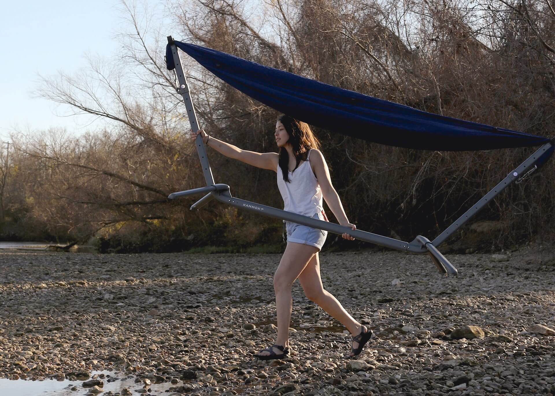 Girl carries a Swiftlet portable hammock stand with midnight blue hammock at chest height walking across a rocky river bed.