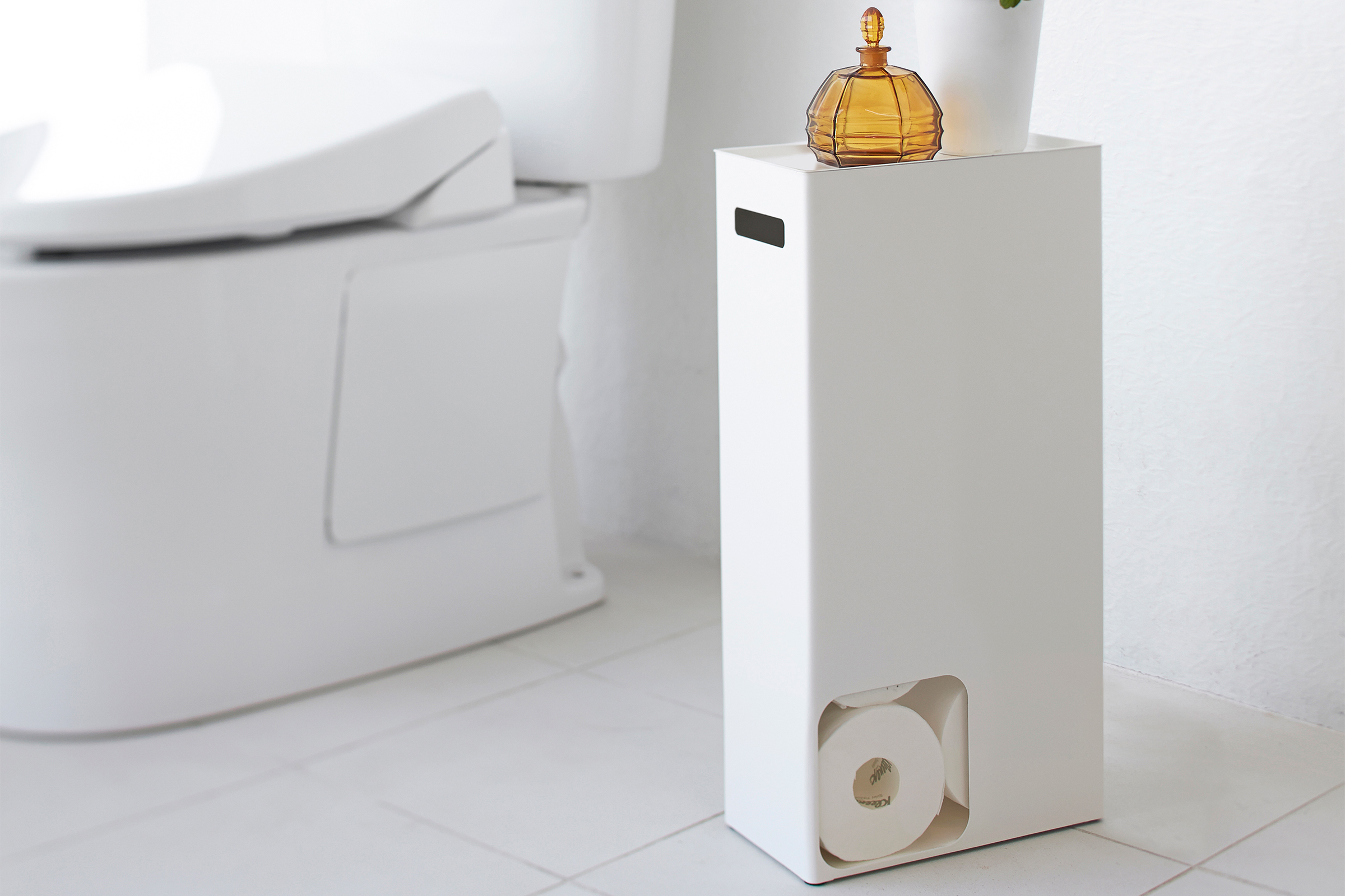 Front view of Yamazaki Home white Toilet Paper Stocker holding toilet paper and home decor items in bathroom.