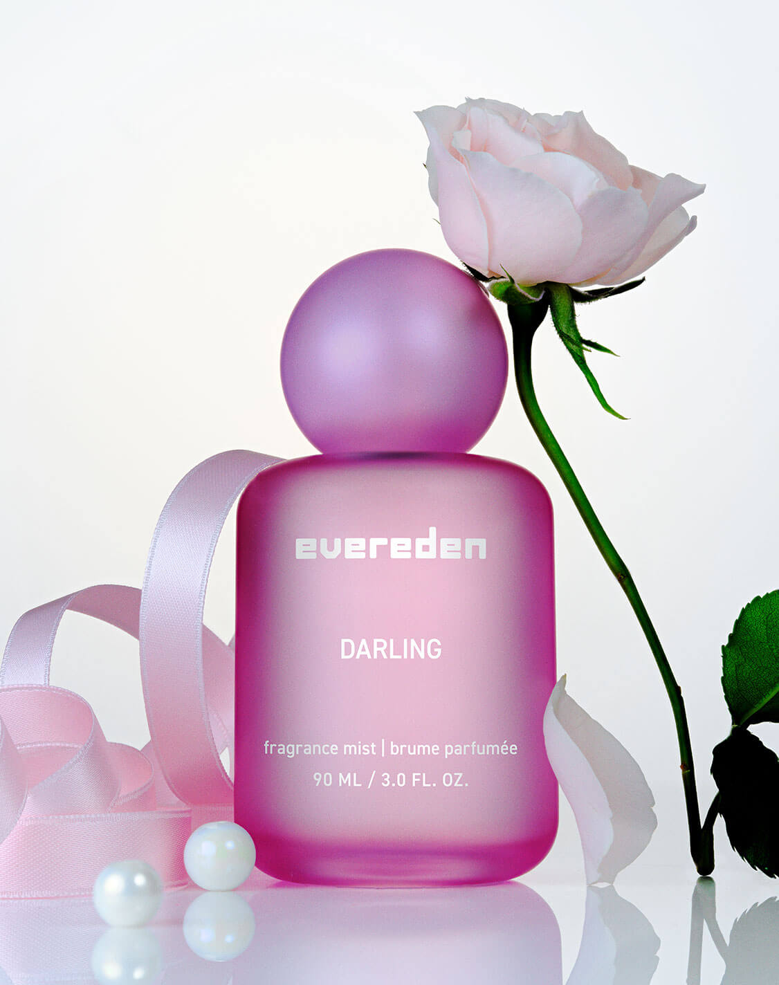 Evereden Darling Fragrance Mist, a frosted pink bottle with a round cap, styled with a pale pink rose, satin ribbon, and pearls on a glossy surface.