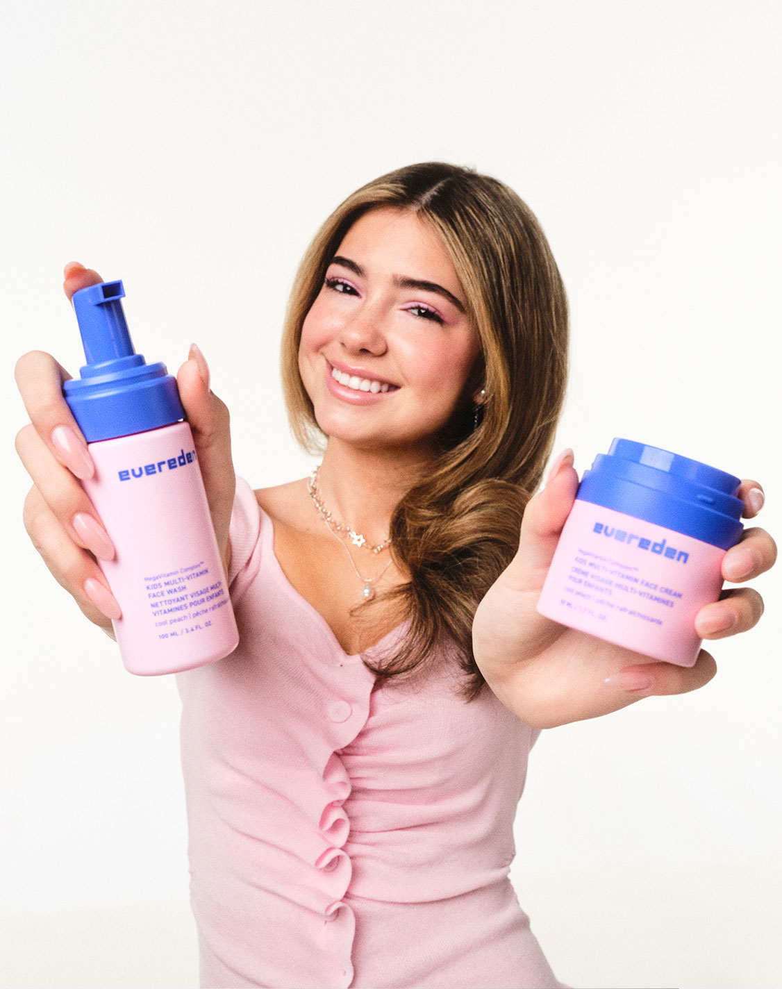 A smiling girl holds out two Evereden products toward the camera — Kids Multi-Vitamin Face Wash in her right hand and Kids Multi-Vitamin Face Cream in her left.