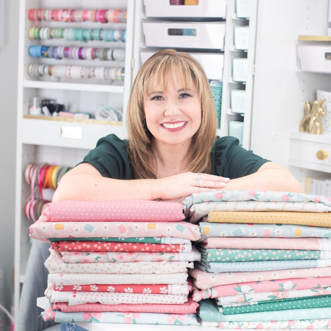 a smiling woman is posing with a tall pile of colorful folded fabrics.