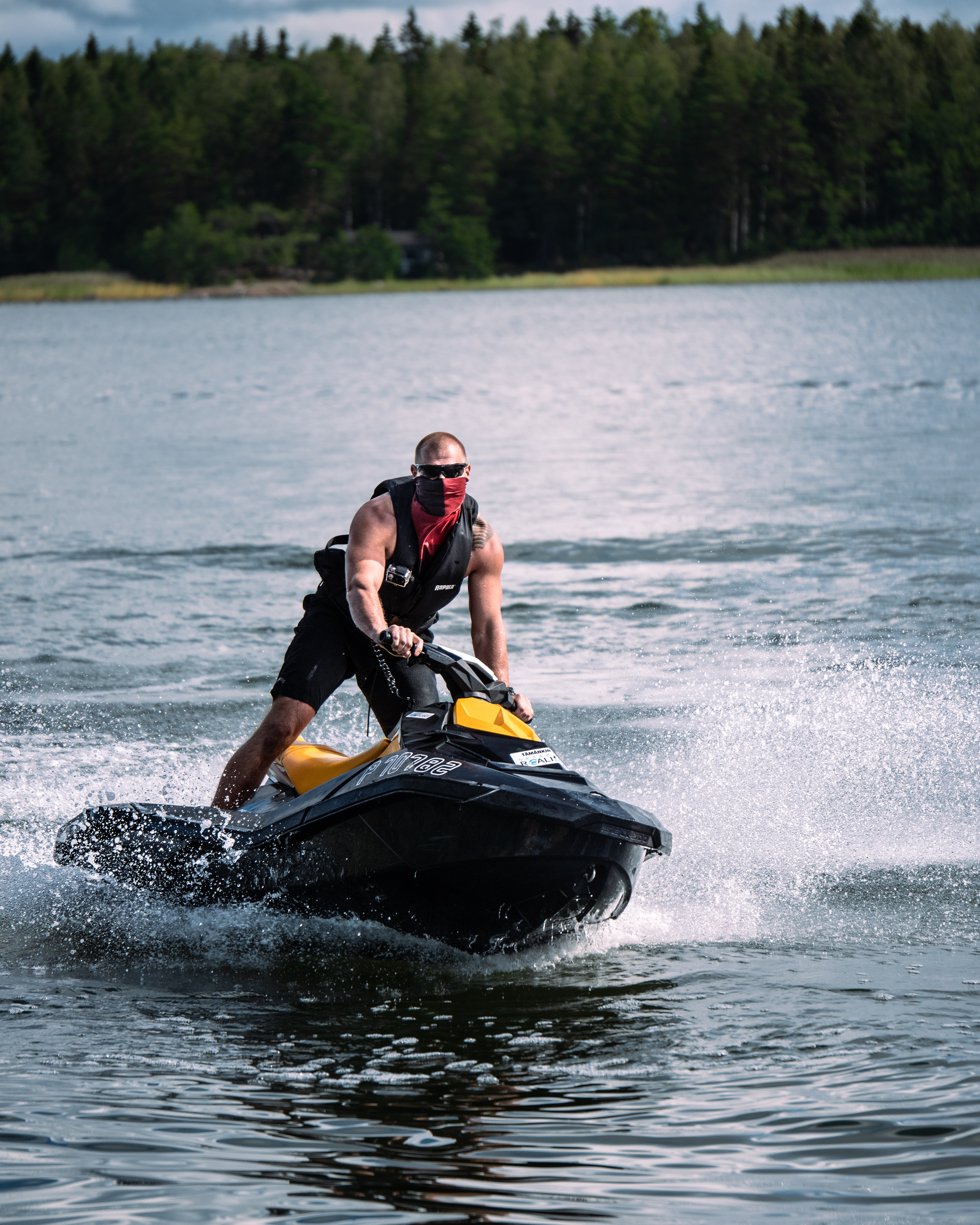 Muscular man riding a jet ski. Wearing a red bandana, sunglasses, a life west and shorts. He is in a lake.