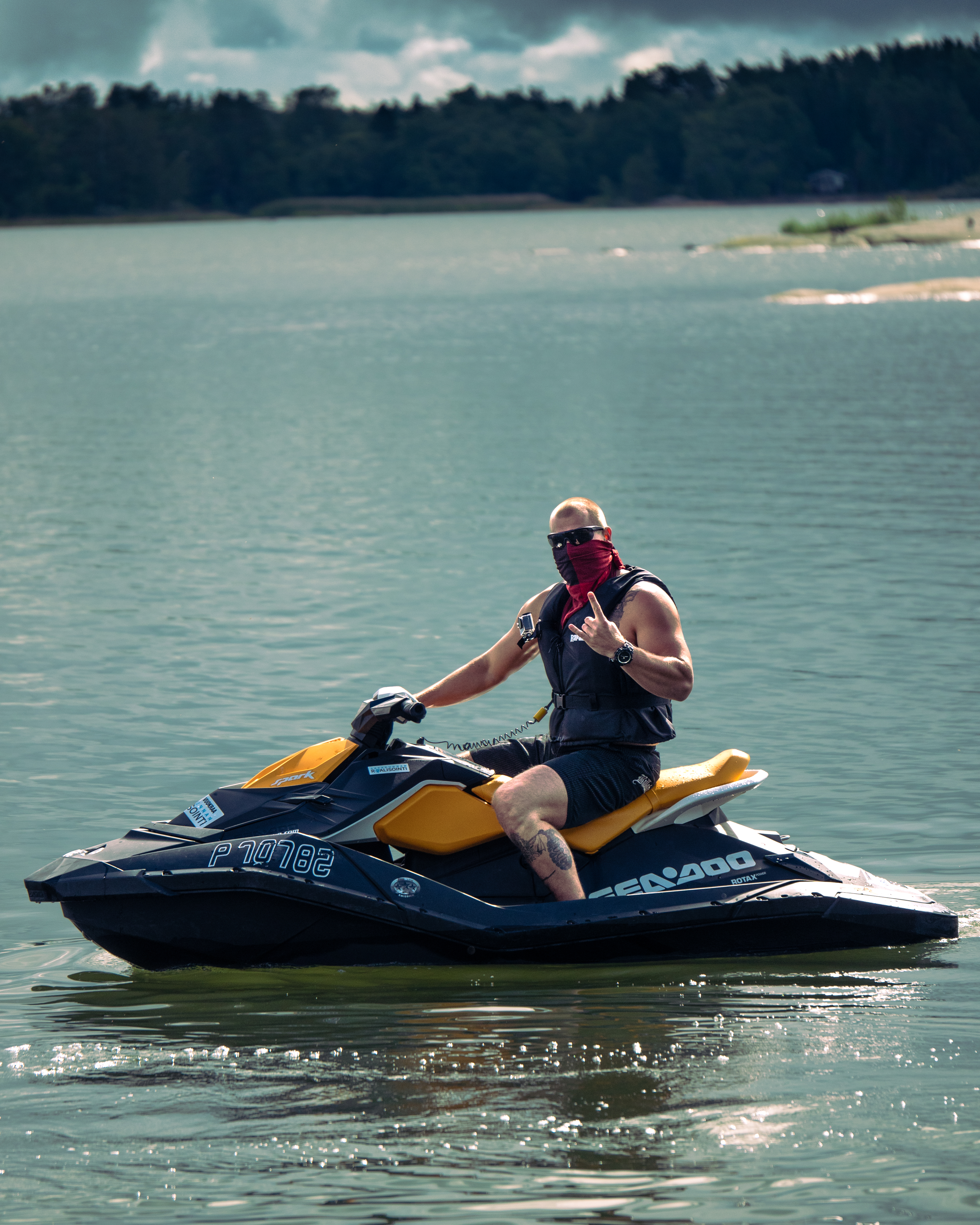 Muscular man on a jet ski. Wearing a red bandana, sunglasses, a life west and shorts. He is in a lake.
