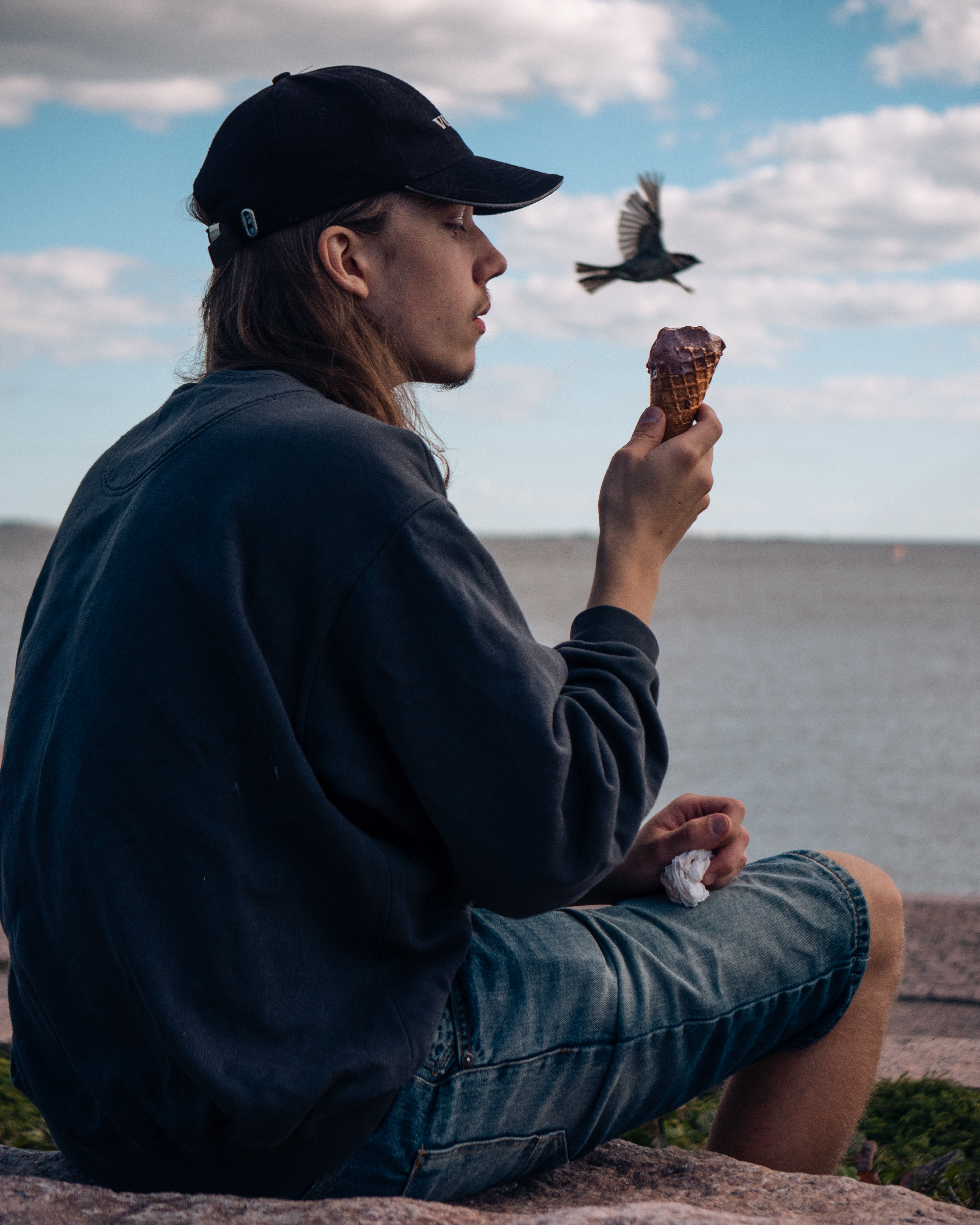 Man with long hair eating ice cream outside by the sea. A bird is flying by. He is wearing pullover and jeans.