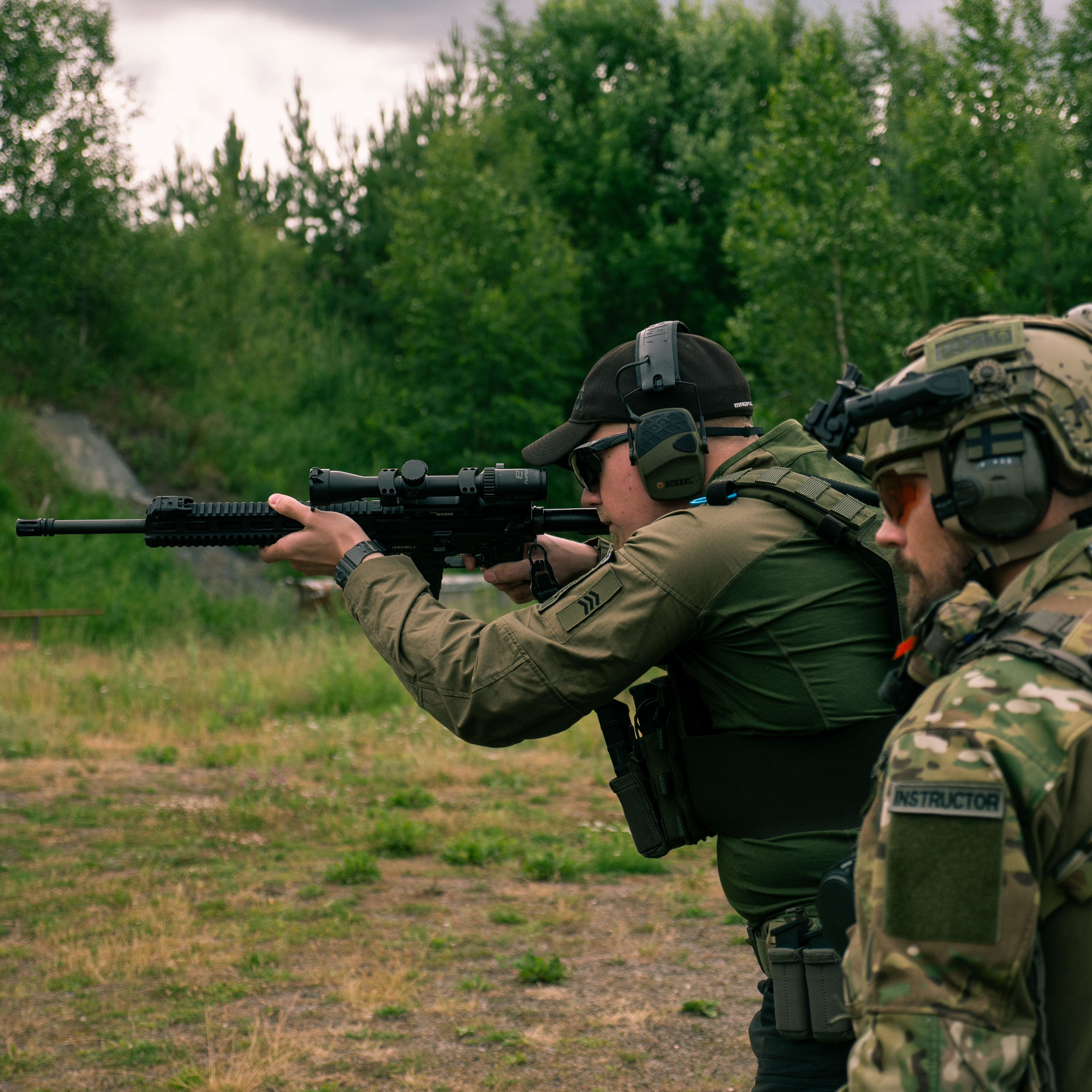 Man shooting a rifle. The rifle has a scope on it. The man is wearing a jacket. He is on a shooting range.  A instructor is following him