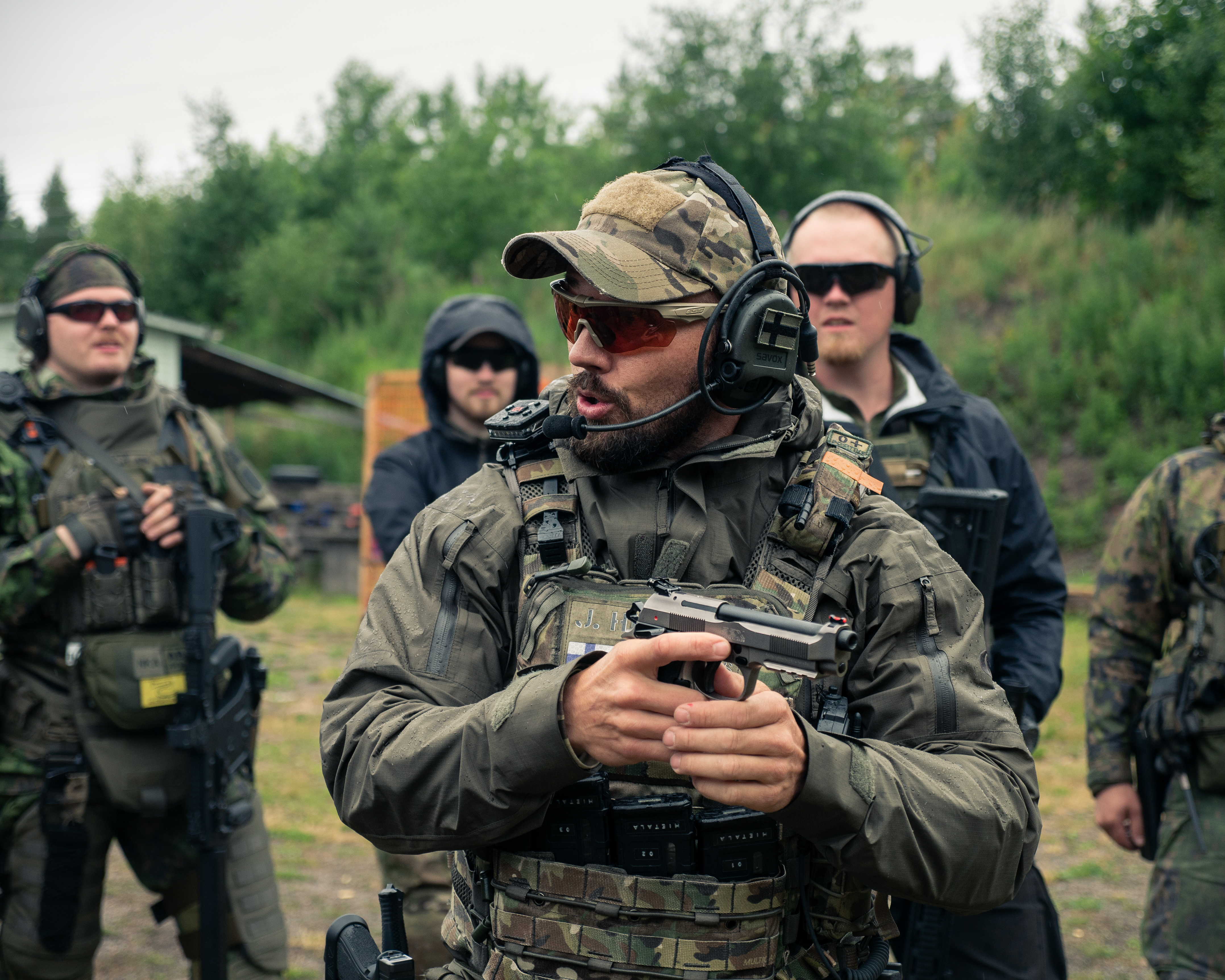 A man Jere Hietala is standing on a shooting range. He is wearing earpro, sunglasses, a cap, jacket and a plate carrier. He has a beard.  He has a pistol. Men are standing around him.