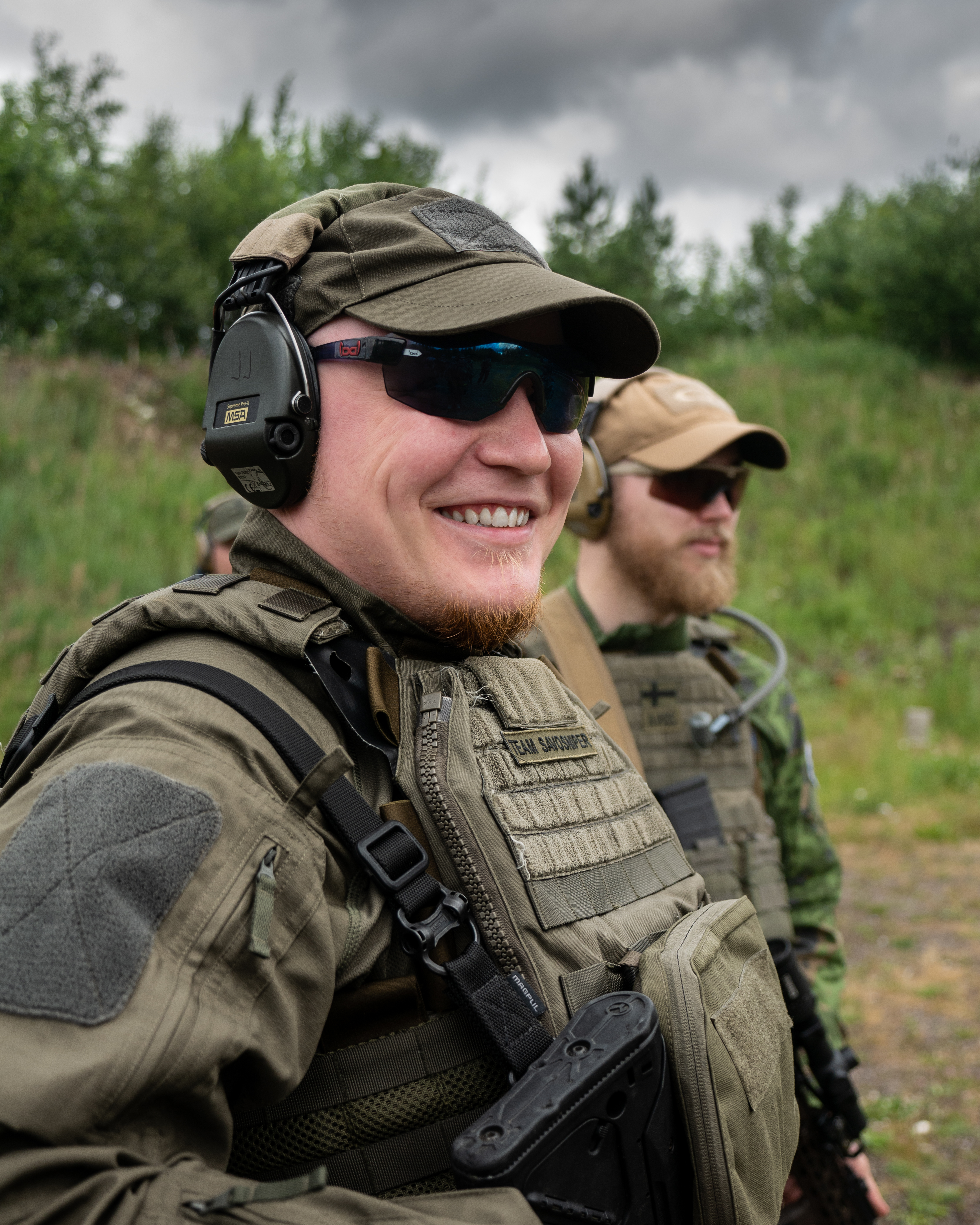 A man with sunglasses is smiling. He is wearing uf pro tactical clothing. He has a rifle AR10. There is man behind him. They are on a shooting range.