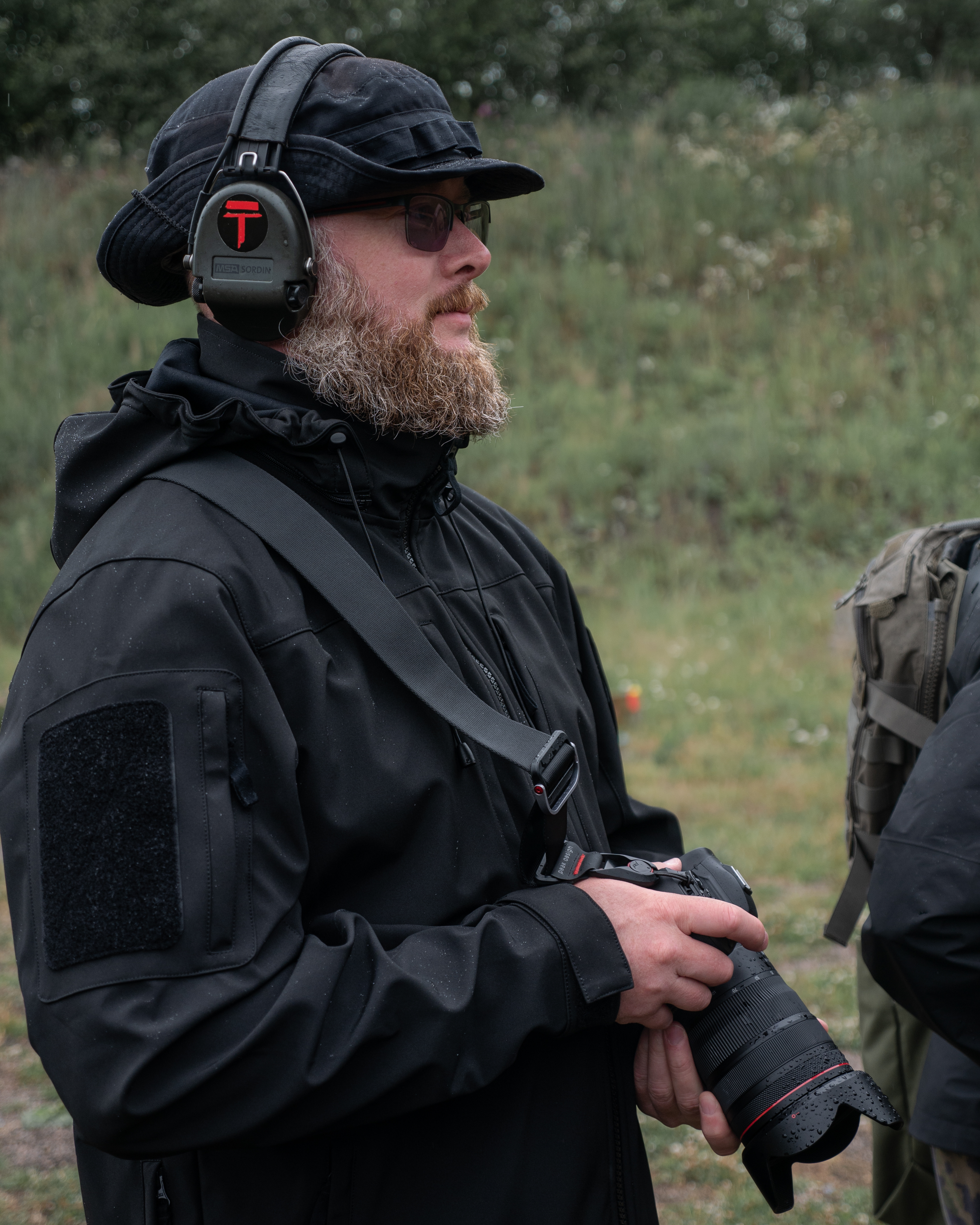 A man with camera. He has a boonie hat, headphones and a black rain jacket. He has a beard and glasses.