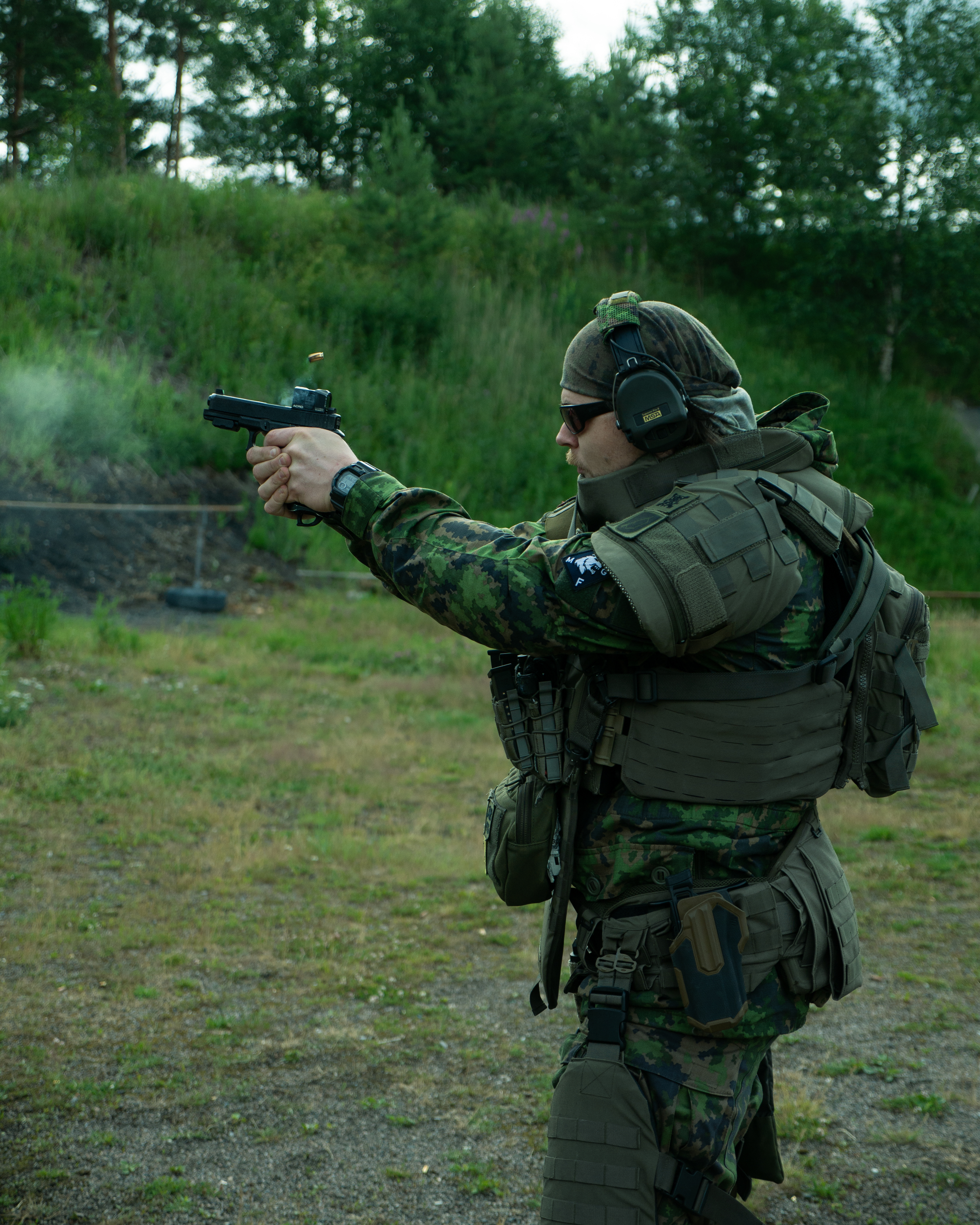 Man shooting a pistol. The pistol has a red dot on it. The man is wearing body armor and a M05 camo jacket. He is on a shooting range.