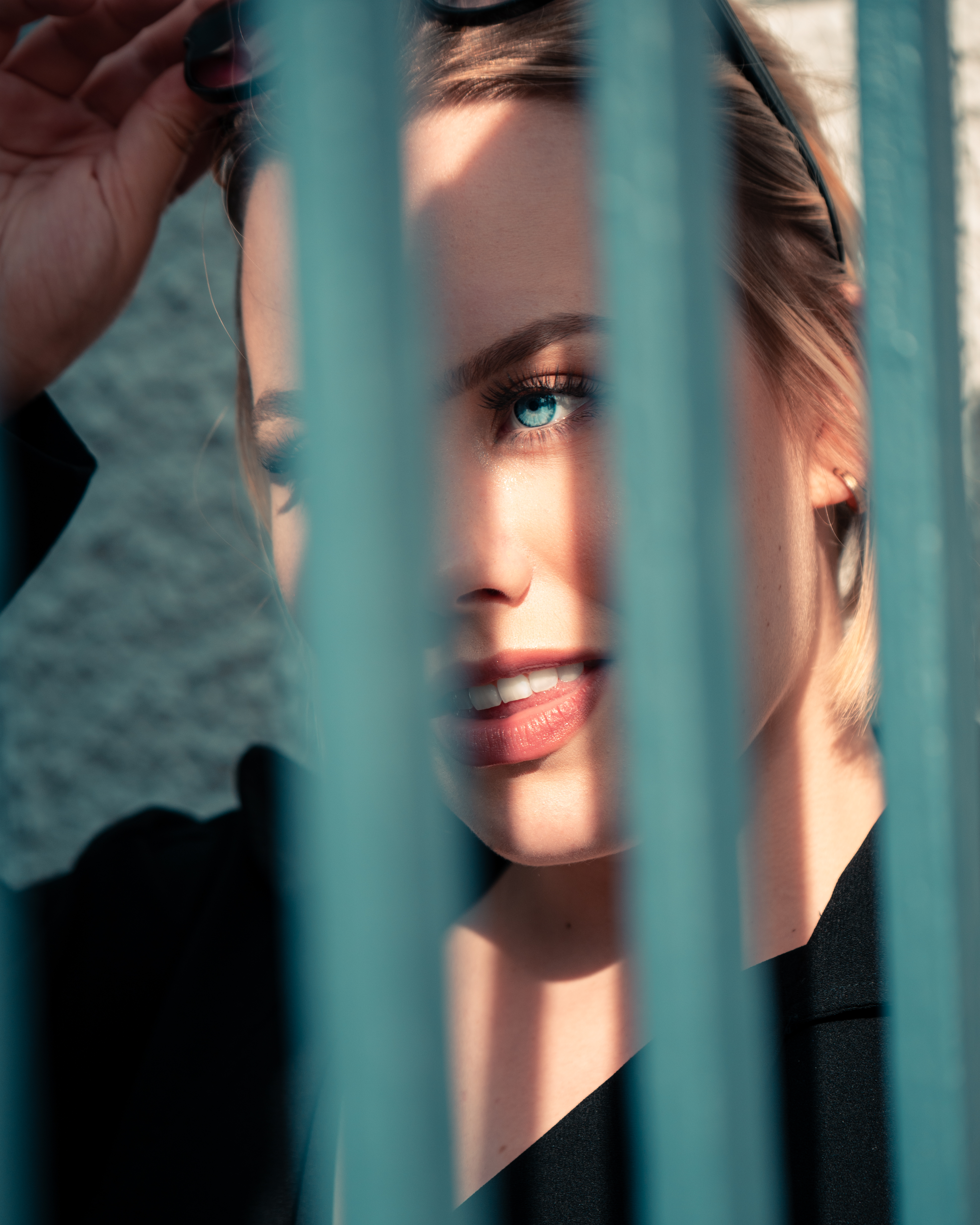 A blonde woman with blue eyes is standing behind bars. Light shines on her face. She is smiling. She has sunglasses on her head and she is holding them with her hand. She is wearing a black coat.