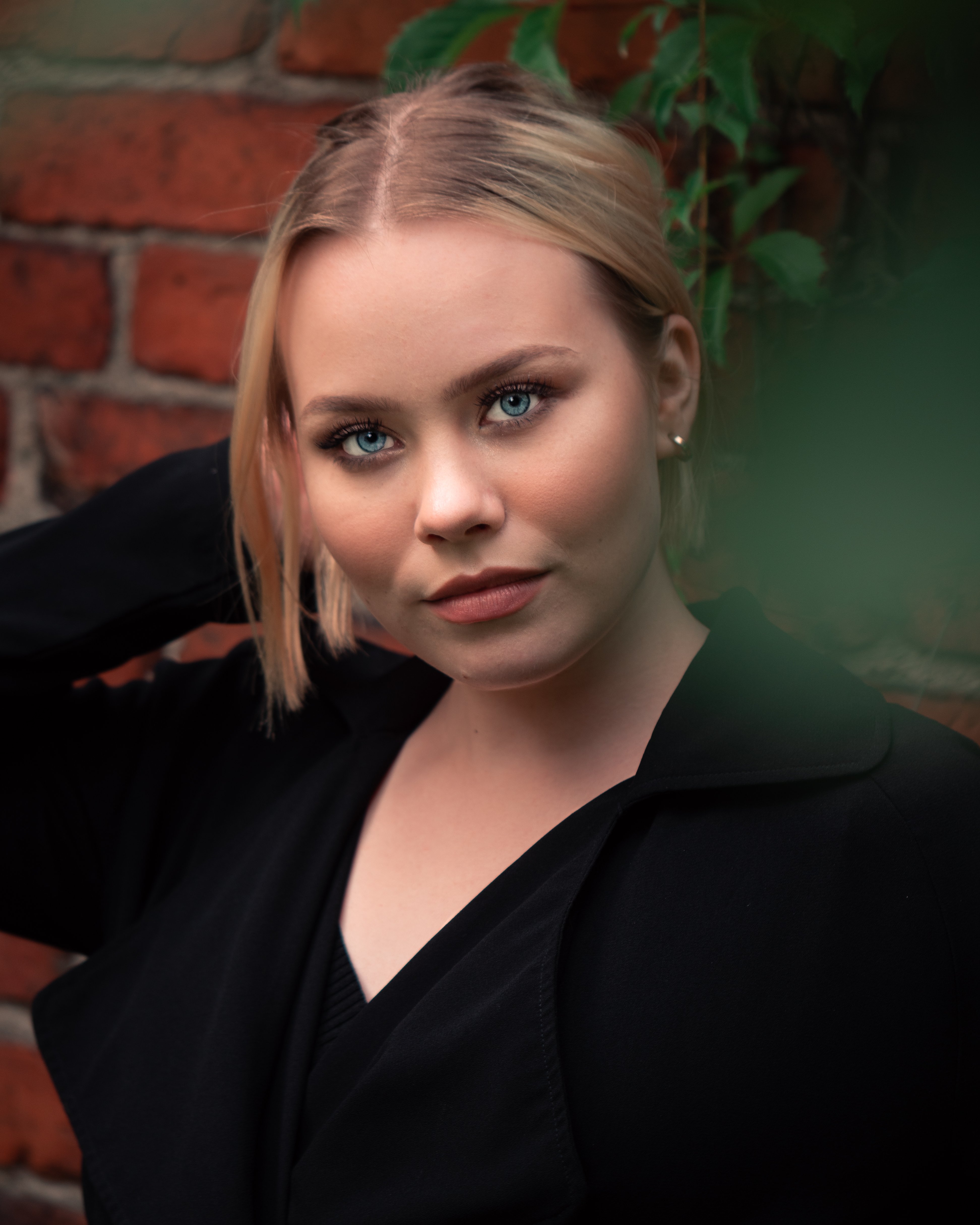 Blonde woman with blue eyes is wearing a black coat and a black blouse. She is standing in front of a brick wall. She has a neutral expression.