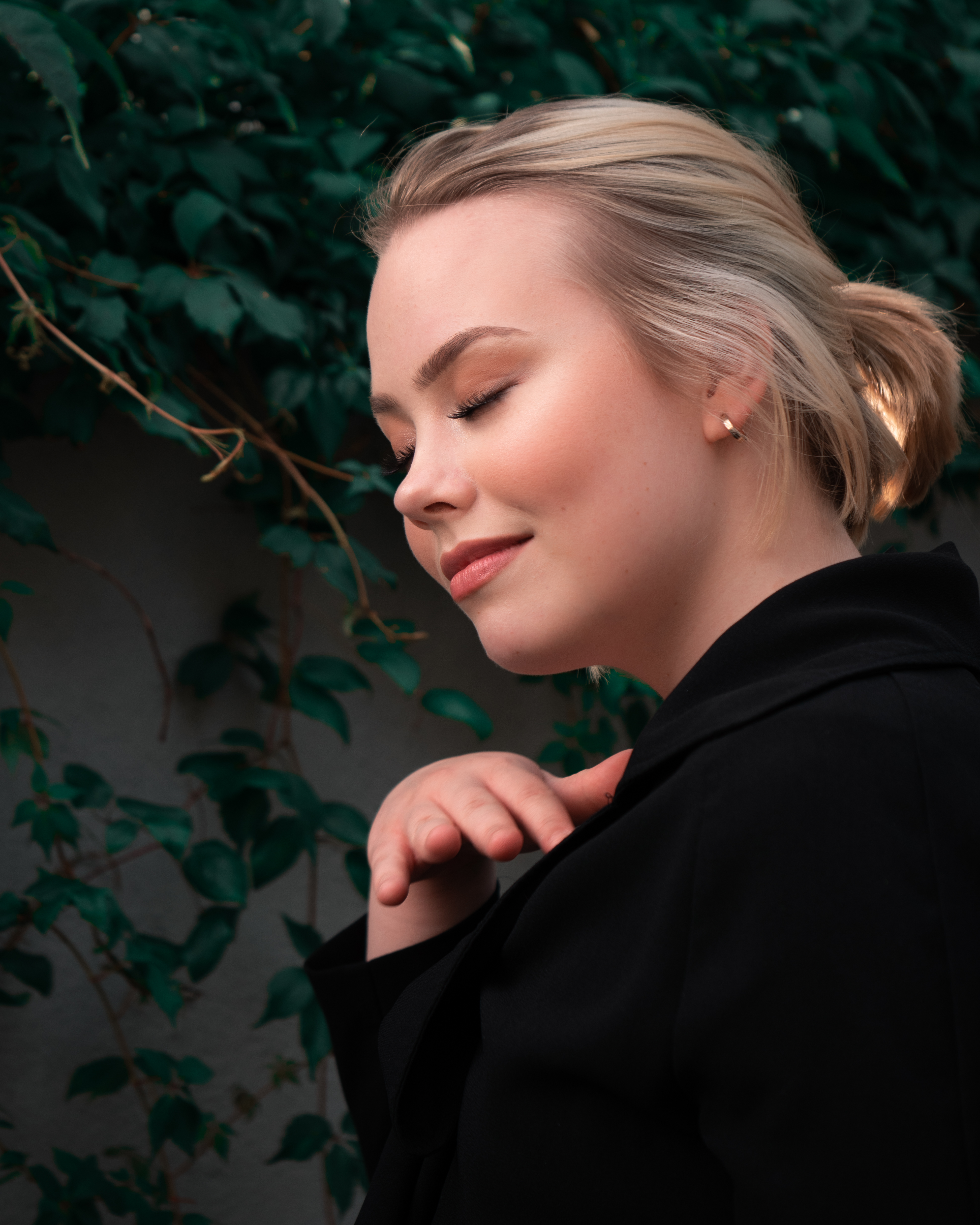 Blonde woman with blue eyes is wearing a black coat and a black blouse. She is standing in front of a wall that has leaves growing on it. She is smiling with her eyes closed.