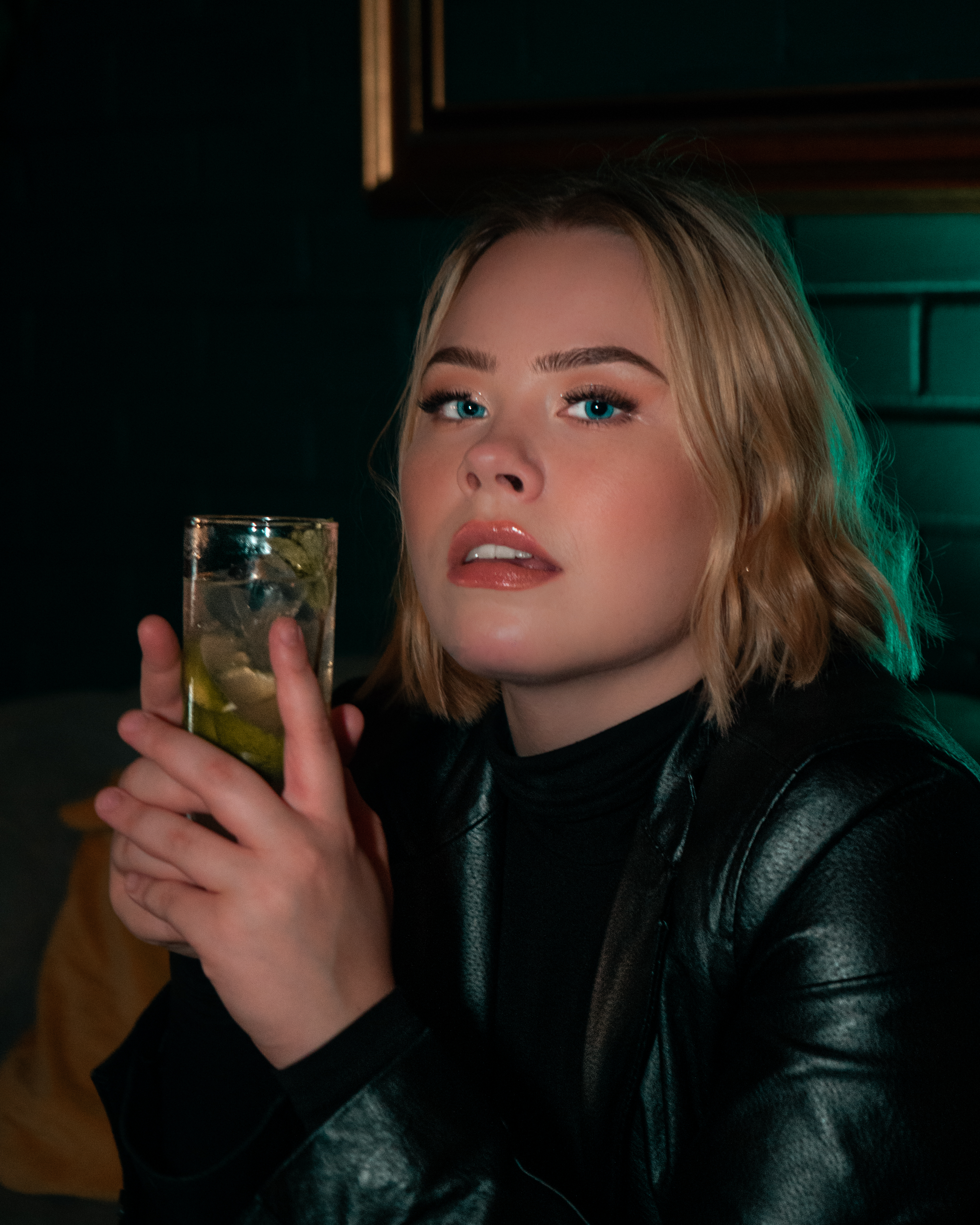 Blonde woman with blue eyes is wearing a leather jacket and holding a Moscow mule drink in her hand. She is smiling while she is sitting in a bar on a sofa.