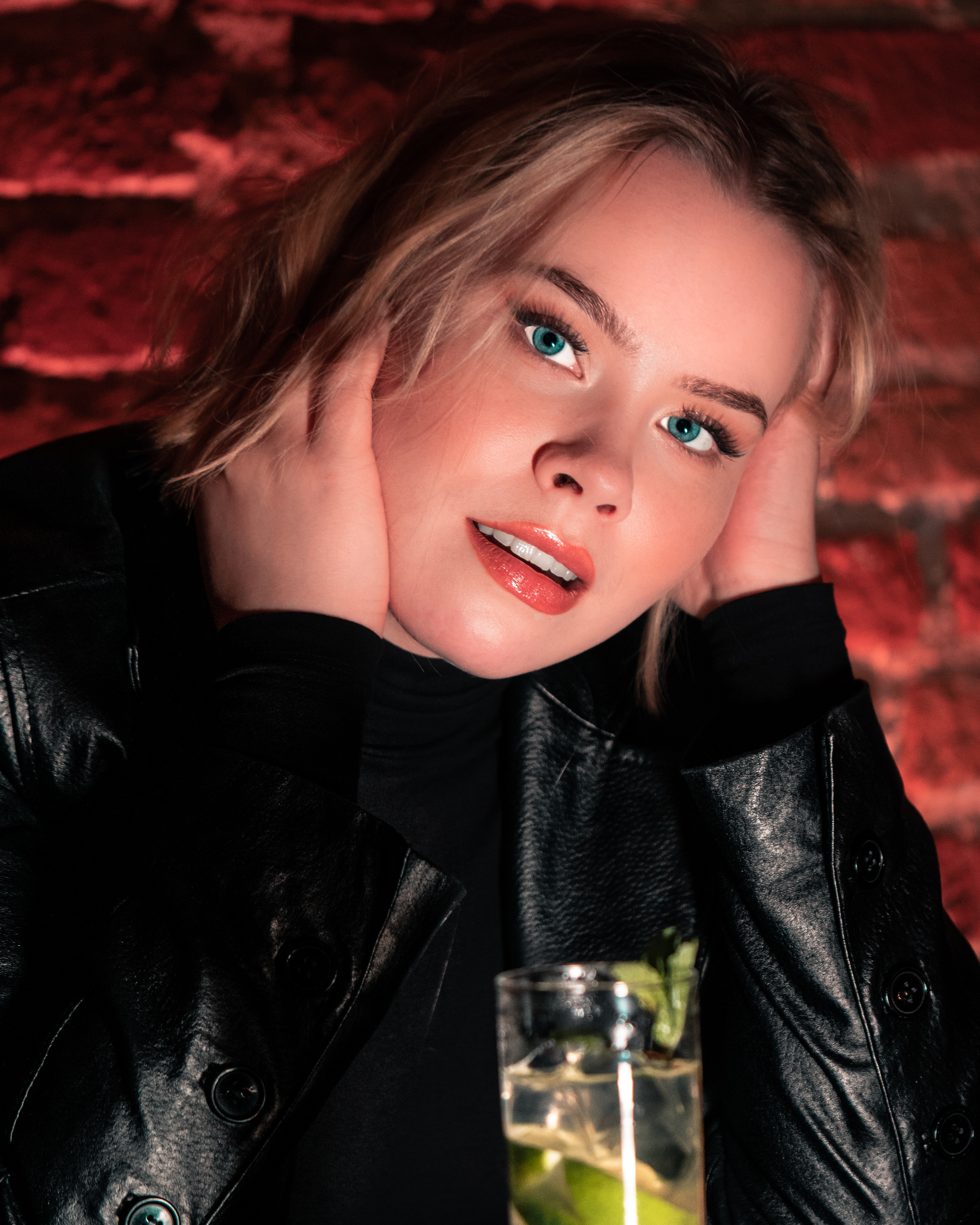 Blonde woman with blue eyes is wearing a leather jacket. A Moscow mule drink is standing on the table in front of here. There is a red brickwall behind her. She is smiling while she is sitting in a bar.
