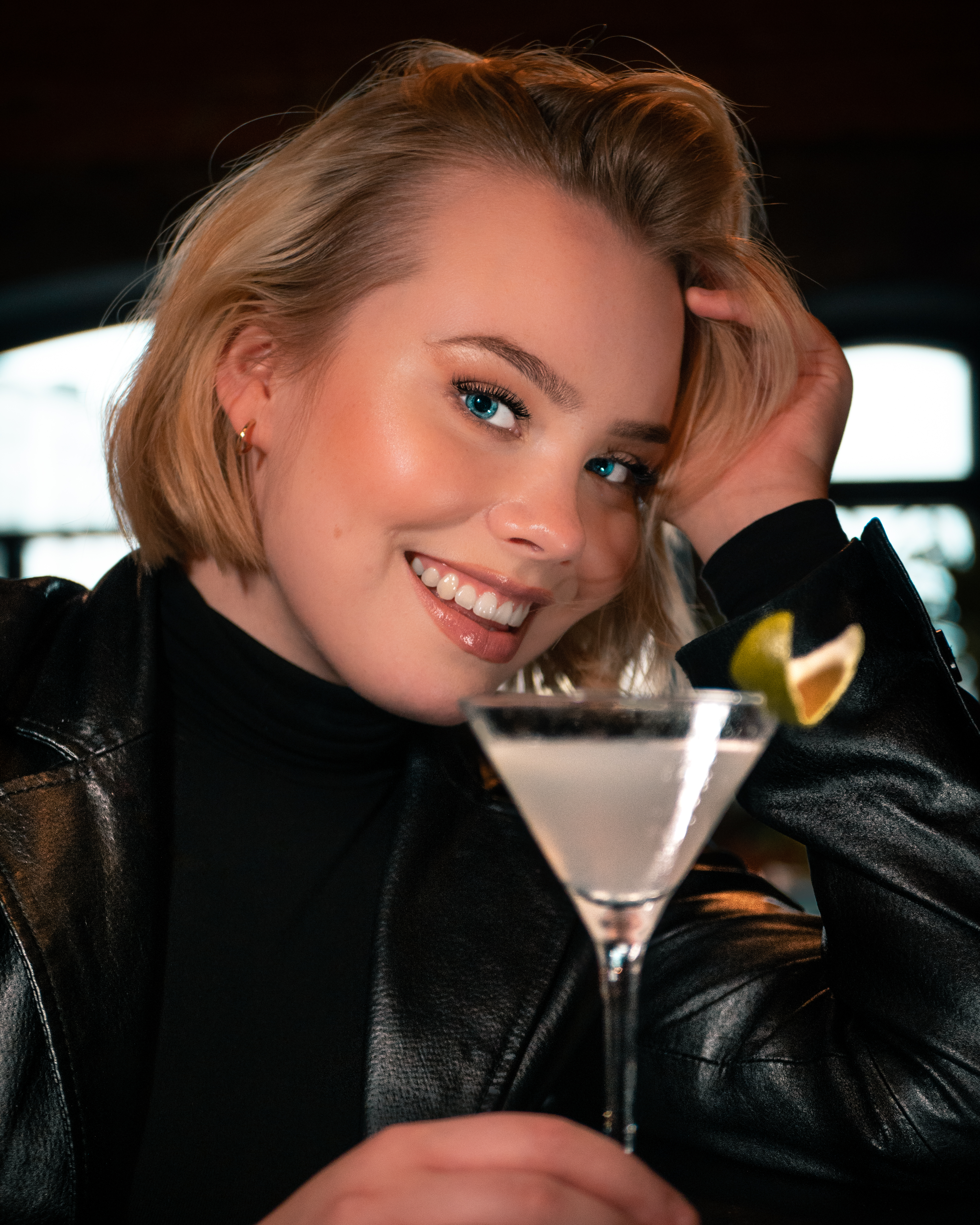 Blonde woman with blue eyes is holding a drink and smiling at the camera. She is wearing a black leather jacket.