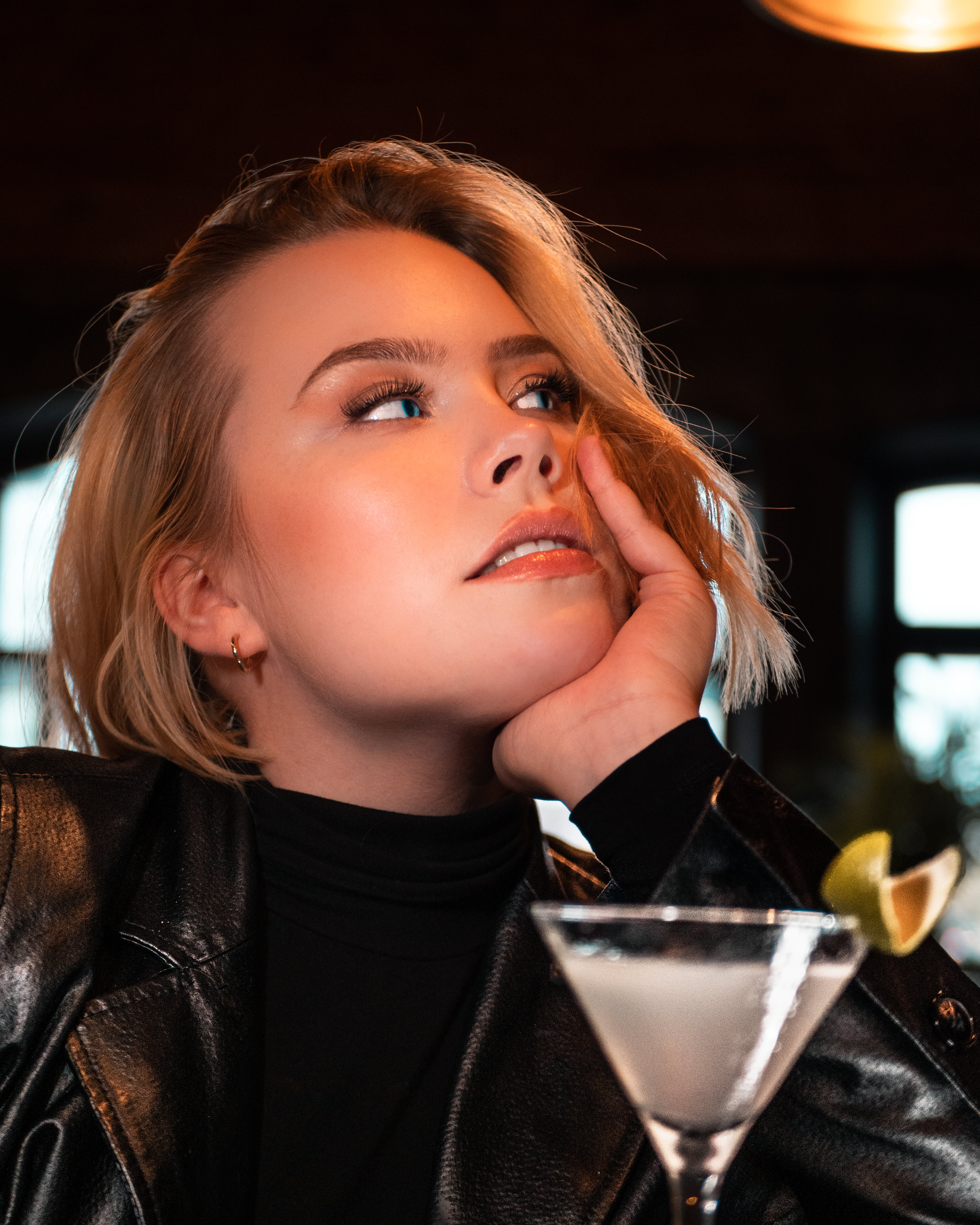 Blonde woman with blue eyes is holding a drink and she is looking away from the camera and seems to be in deep thought. She is wearing a black leather jacket.