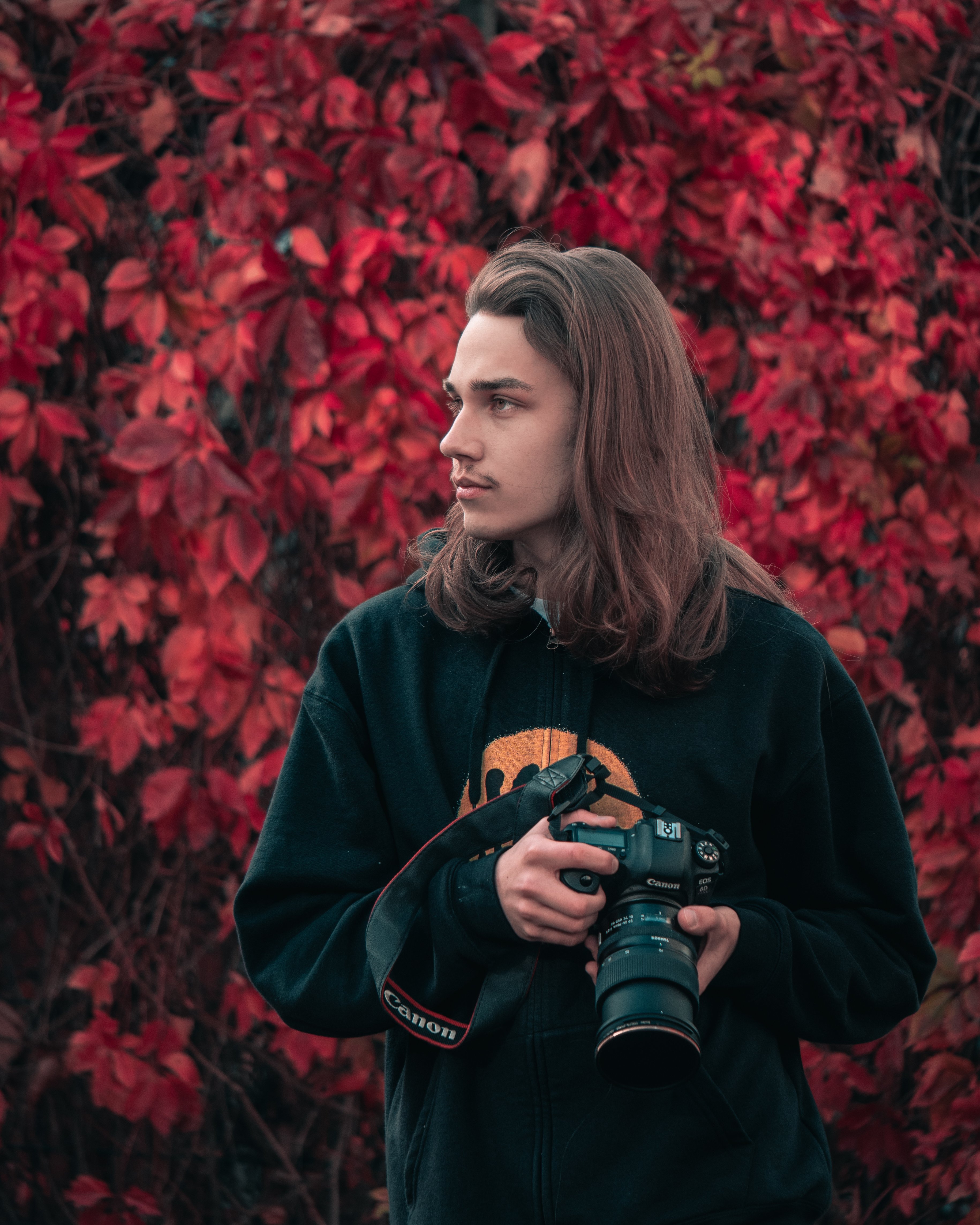 Man with camera standing in front of red leaves. He is wearing a black hoodie with a smile on it. He has long hair.