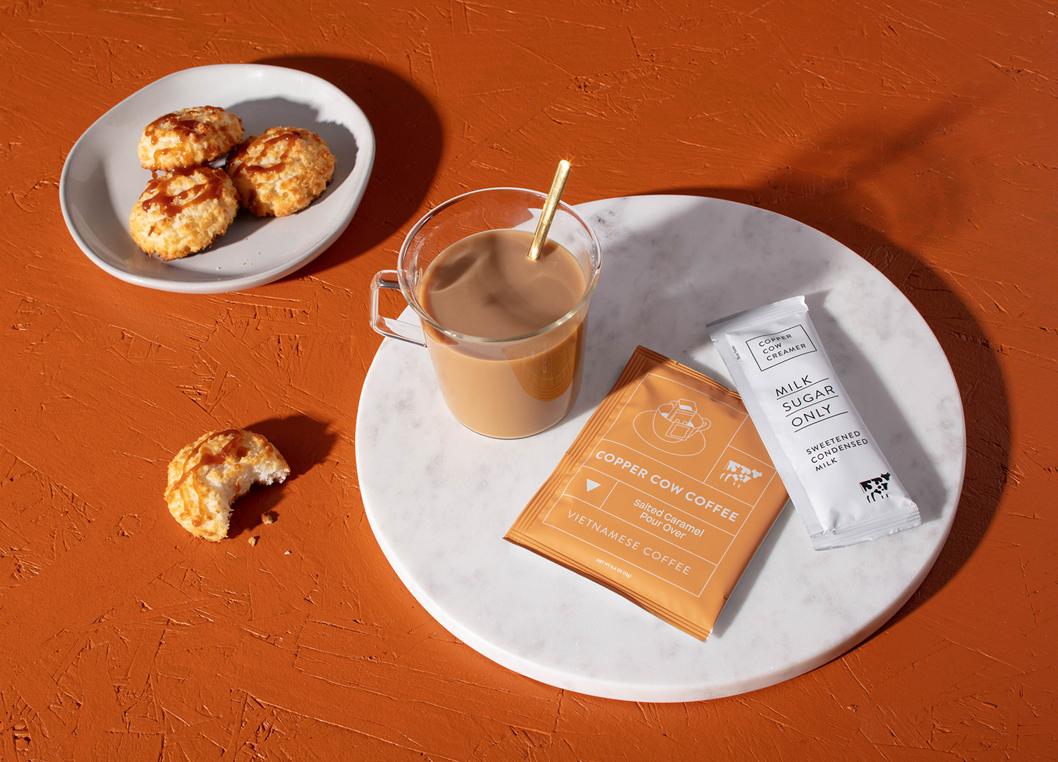 Latte in clear glass mug with spoon sitting next to salted caramel coffee pouch and a creamer packet on a round marble surface next to 1 cookie on the table and 3 cookies on a plate.