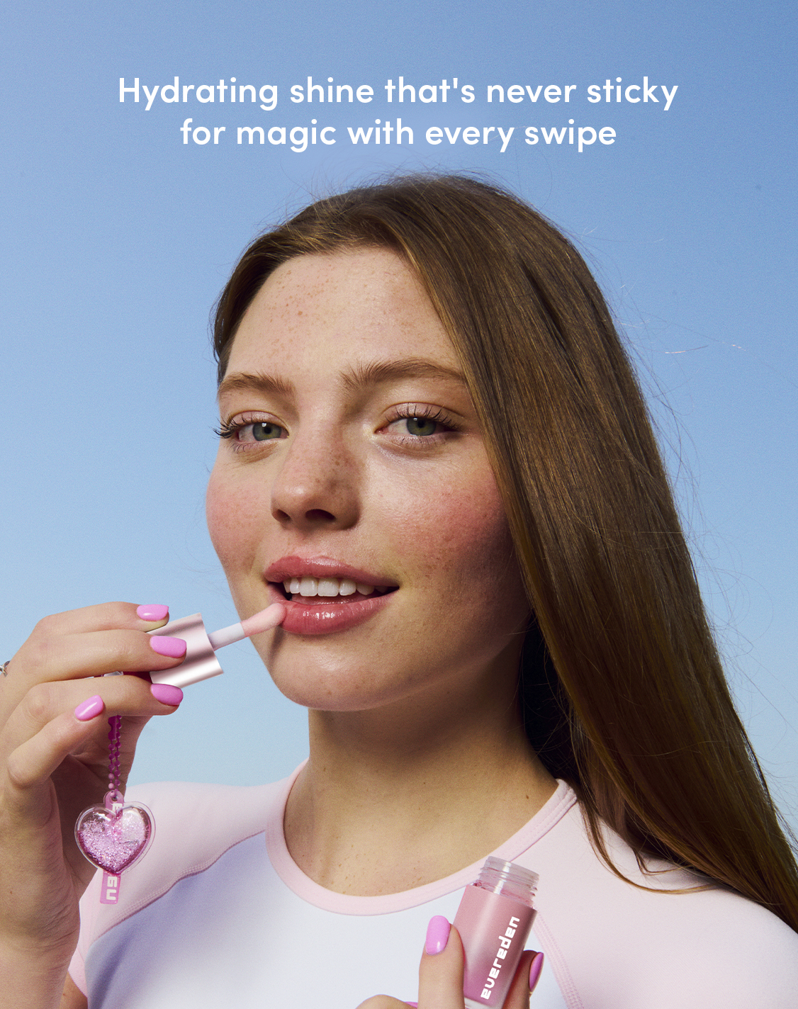 Model applying Evereden Pink Stardust Hydrating Tinted Lip Oil with a doe-foot applicator against a blue background, showcasing its glossy, hydrating finish.