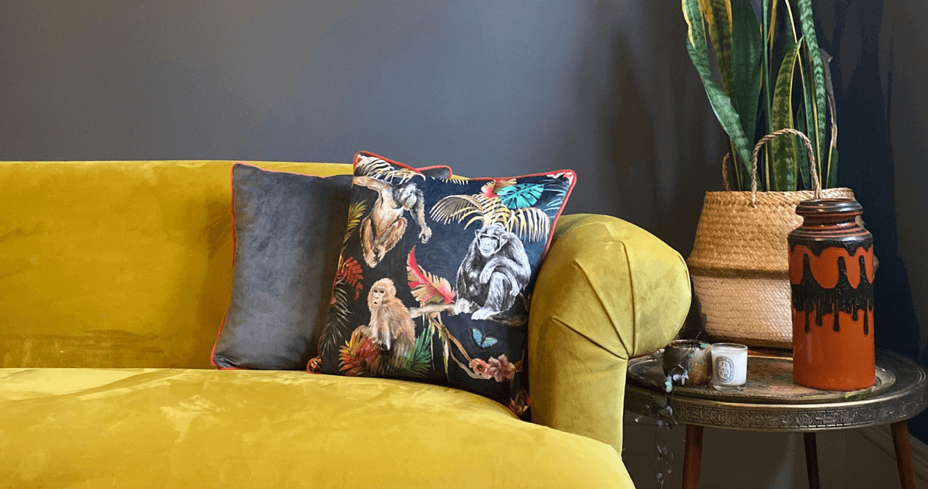 two cushions on a yellow velvet sofa. one is patterned cushion with leaves and monkeys with coral piping and the other is a plain grey cushion with coral piping.