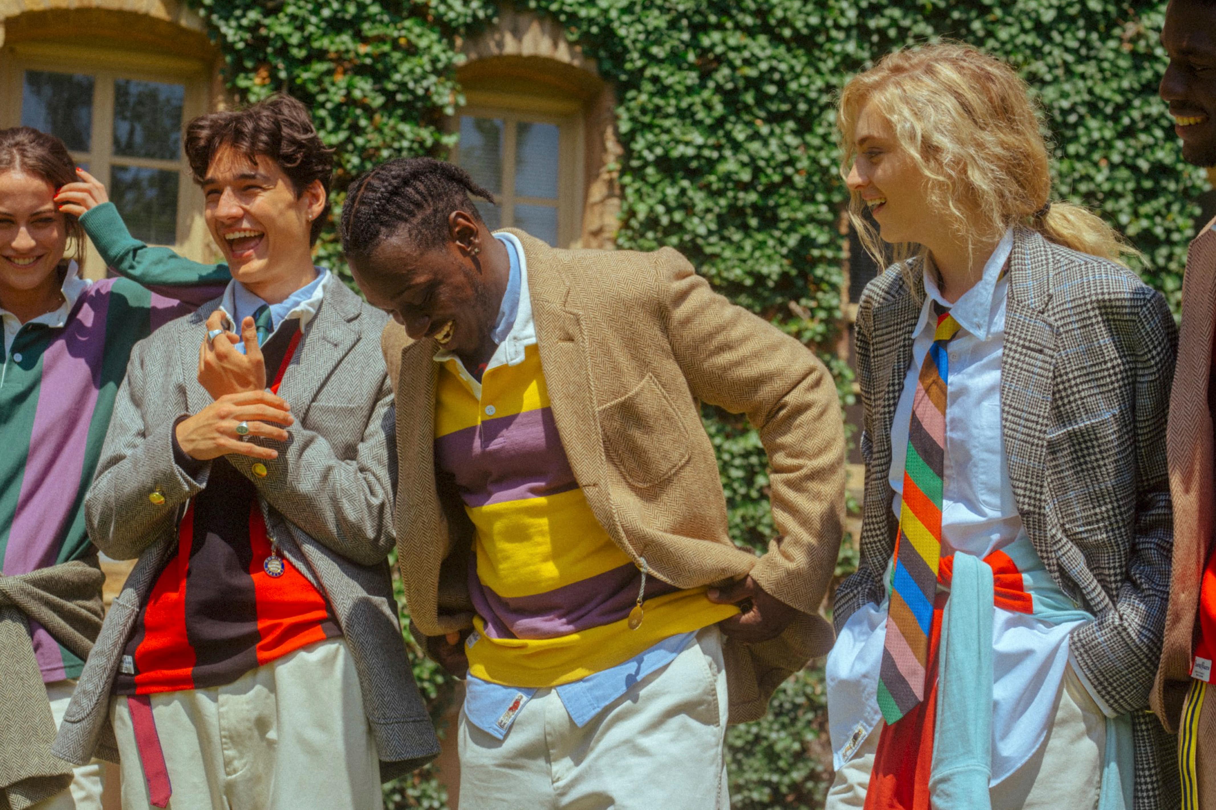 Male model wearing the Vertical Stripe Rugby in black and red. Male model wearing the Horizontal Stripe Rugby in purple and gold. Female model wearing the Oxford Button-Down in blue and the Prince of Wales Jacket
